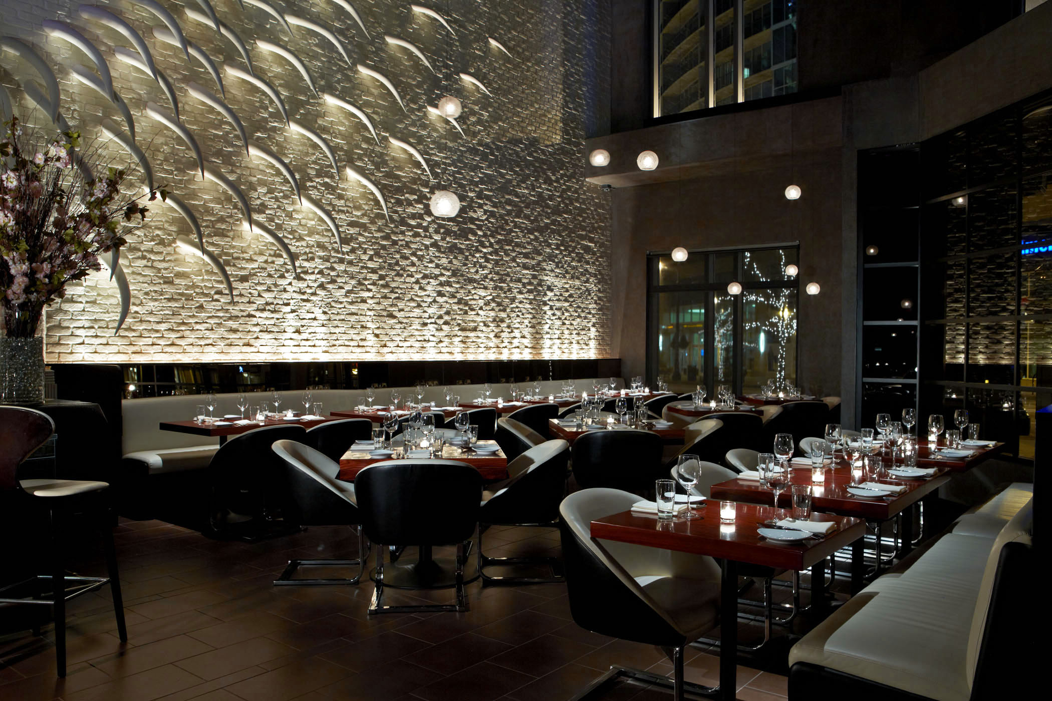 Interior of STK Atlanta restaurant with a textured back wall featuring a repeating pattern of white. The dining area includes dark wooden tables set with glasses and plates, surrounded by a mix of white chairs and curved white banquettes.