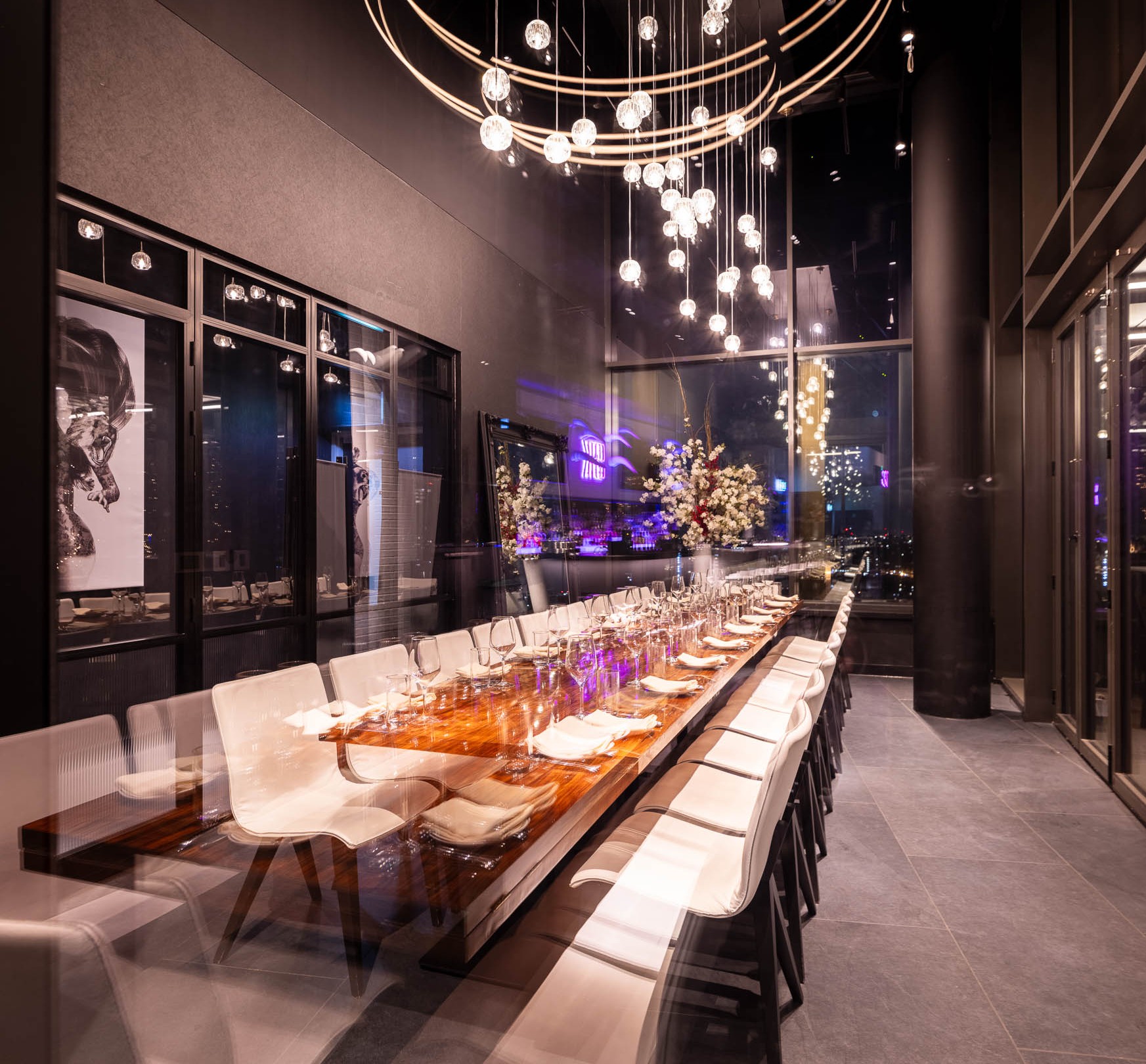 Interior of STK Stratford; a long row of tables and white seats in a soothingly lit room with high ceilings, chandeliers, and hanging artwork.
