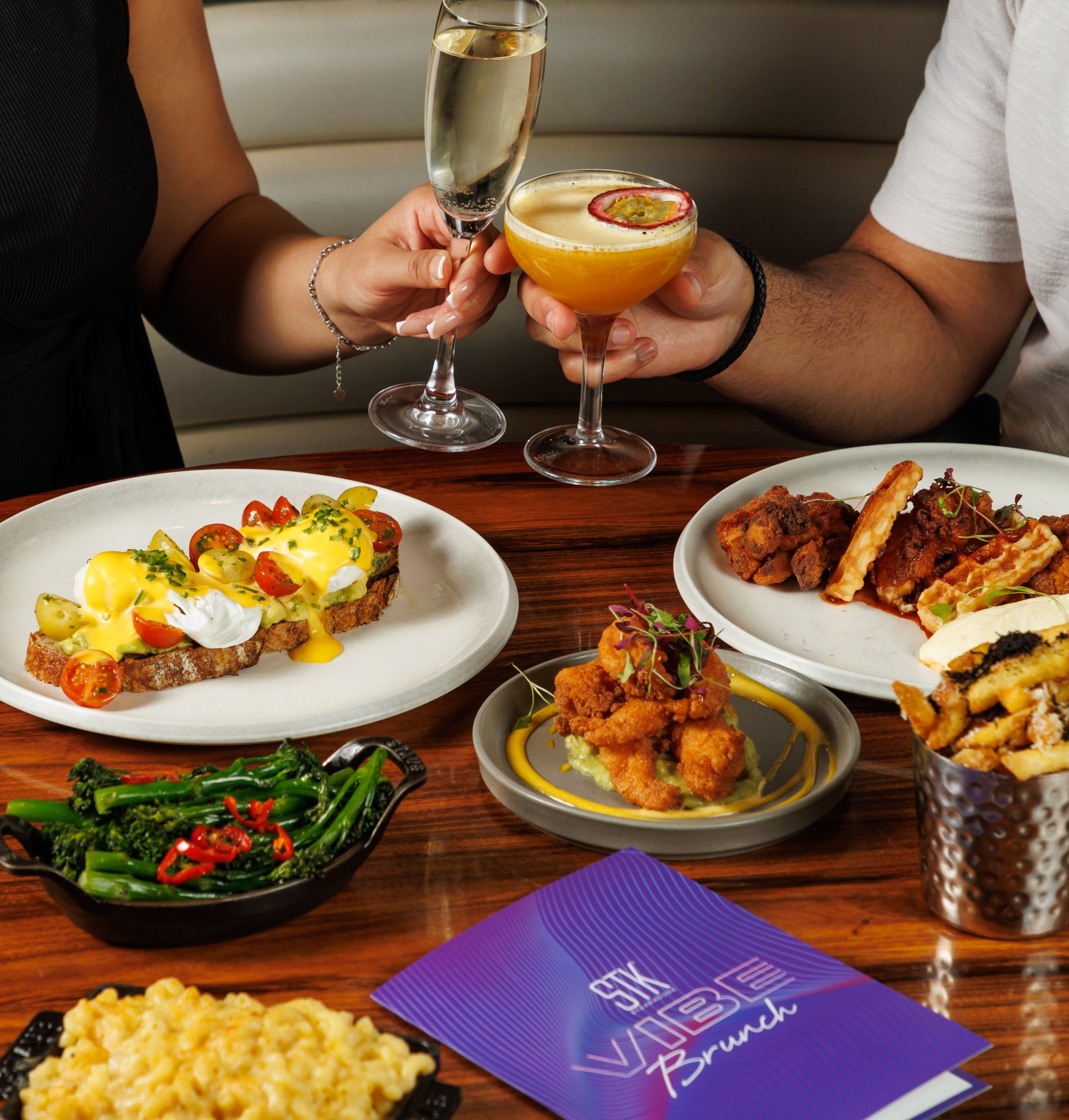 Brunch table spread with eggs Benedict toast, fried chicken and waffles, sides, and a purple “STK Vibe Brunch” menu, while two people toast with champagne and a passion fruit cocktail.
