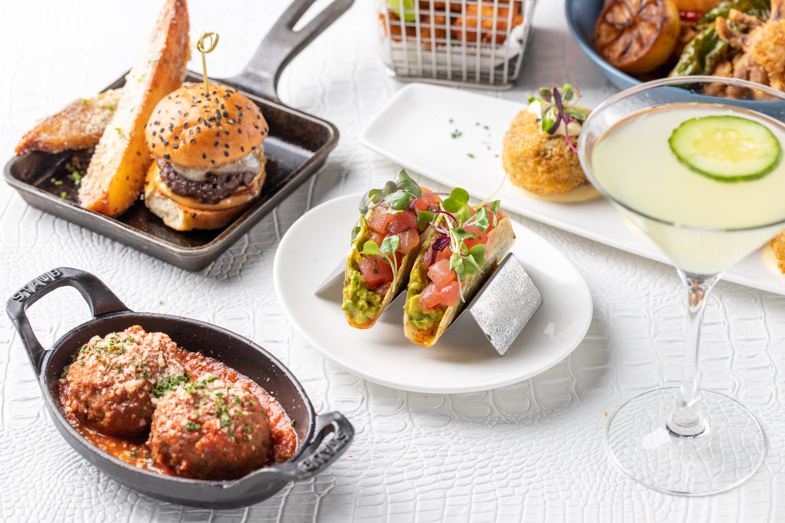 A table laid with various delicious dishes and a cocktail. Including a burger, tacos, meatballs in sauce and a margarita