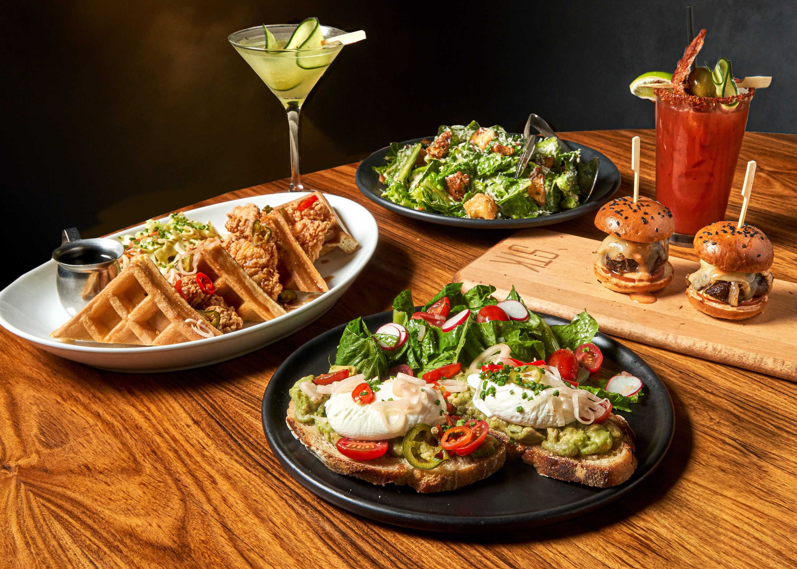 A table laid with delicious dishes and cocktails. Including fired chicken and waffles, mini burgers two colourful cocktails