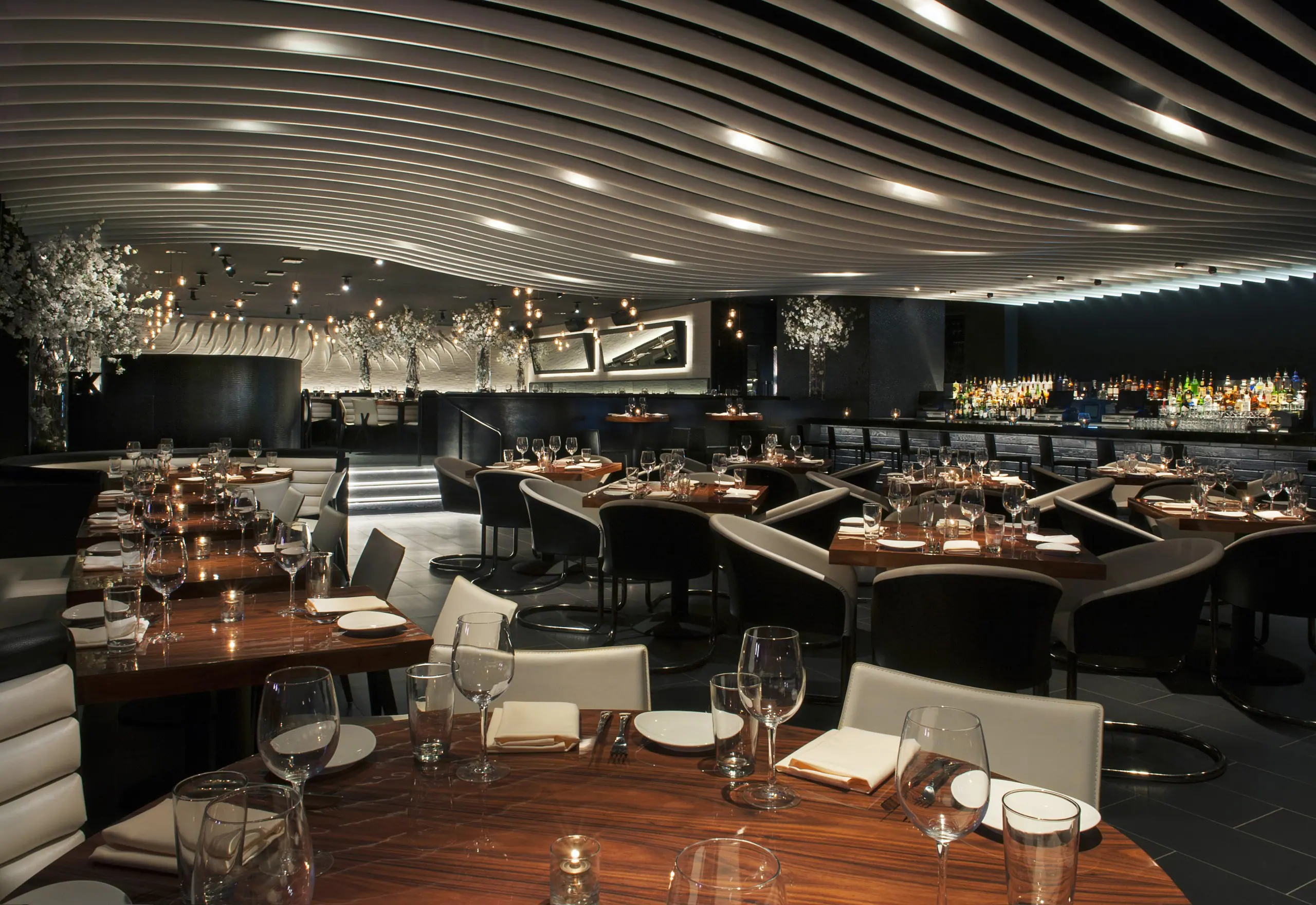 Interior of STK Orlando with a dramatic ceiling installation of curved, wave like panels. The dining area features dark wooden tables surrounded by a mix of black chairs and white cushioned seating.