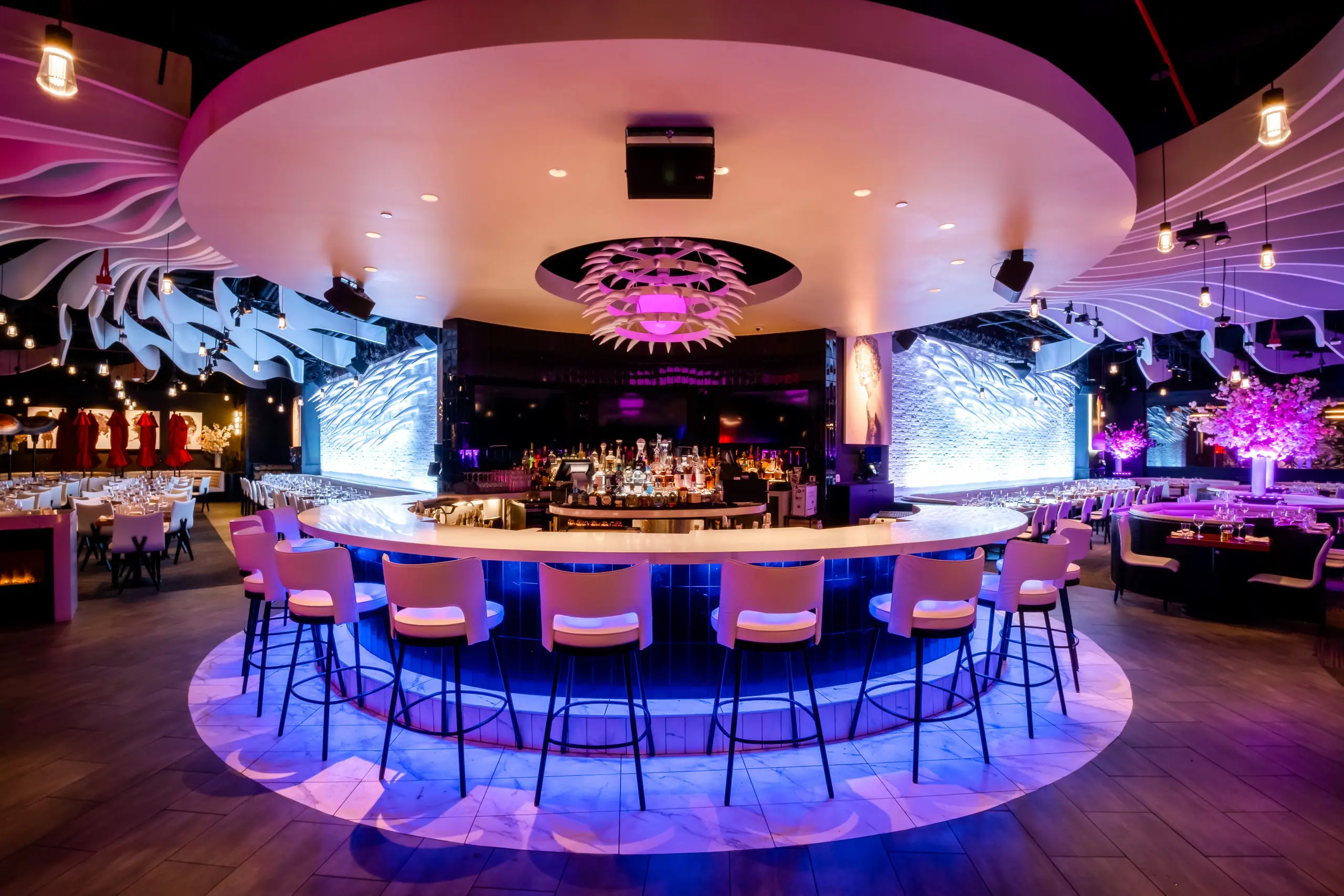 Interior of STK San Francisco bar and lounge with a large circular bar at the centre, surrounded by white barstools. The ceiling features a large, round architectural detail and the space is illuminated by purple and blue accent lights.