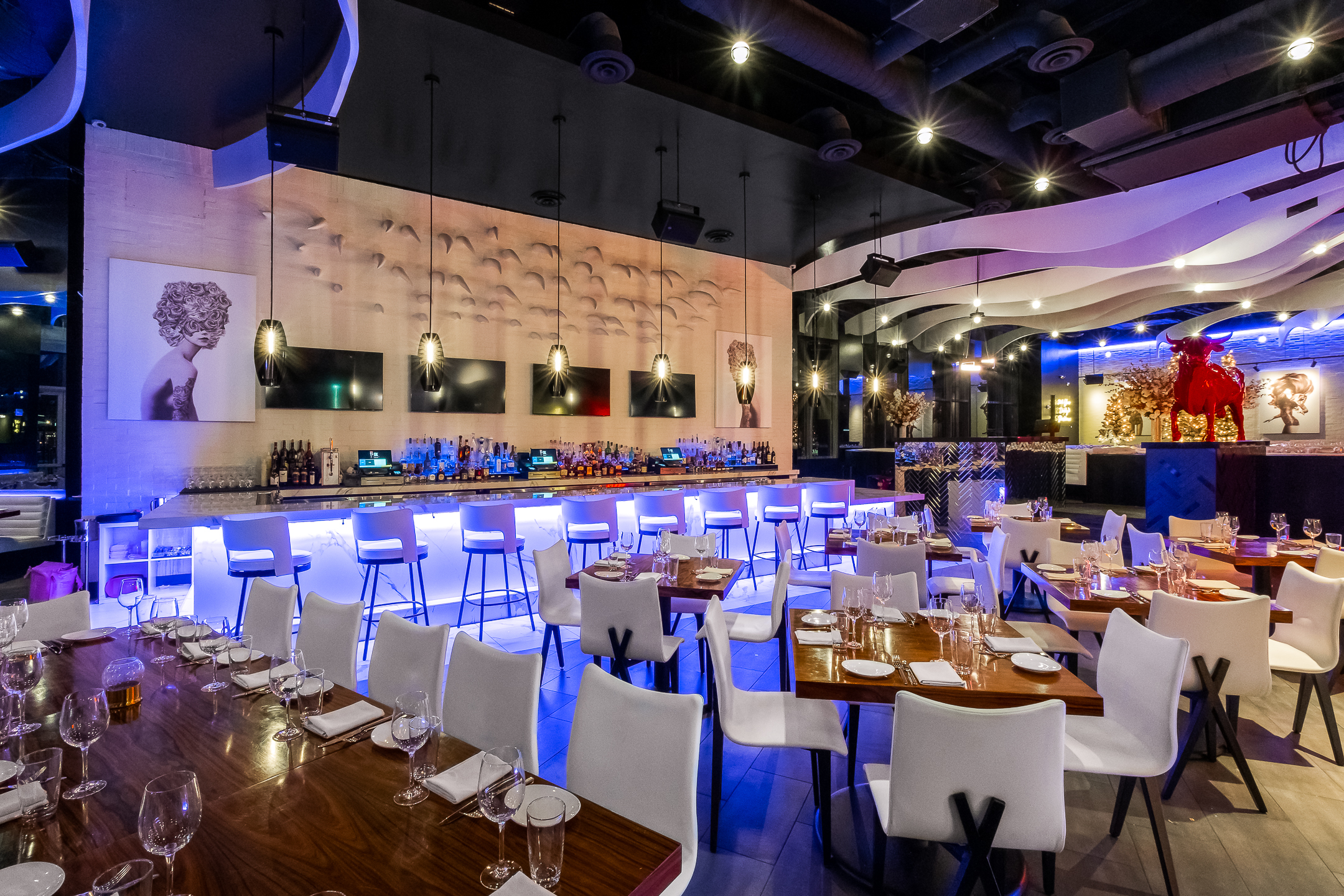 Interior of STK Dallas restaurant, featuring a dining area with fully set tables. The tables are arranged with white chairs, glassware, plates, and folded napkins. A bar in the background is illuminated with blue lighting, lined with high stools and a variety of bottles. The ceiling has wave like panels and pendant lights.