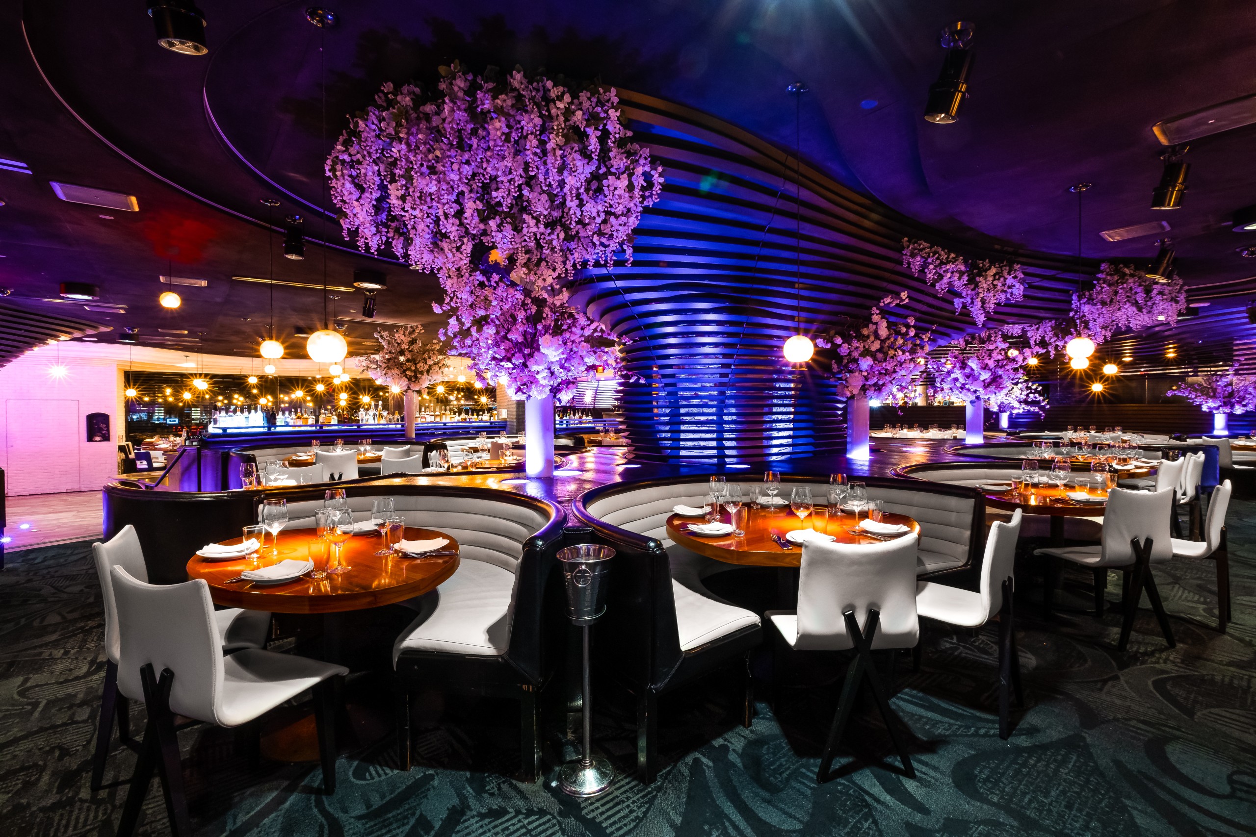 Interior of STK Las Vegas with dark curved booth seating arranged in semi circles around round wooden tables. A blue textured wall and cascading floral ceiling details form the backdrop.
