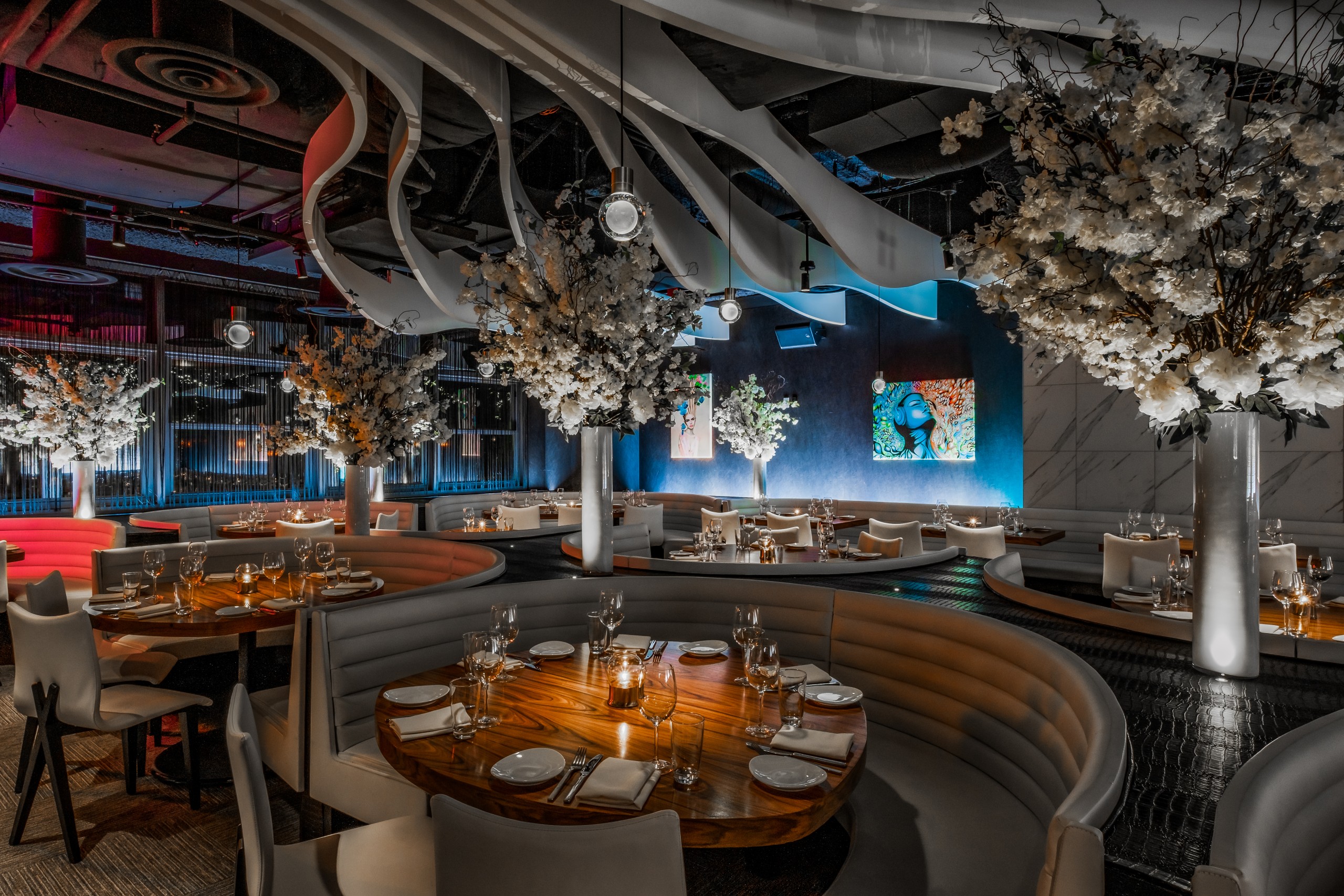 STK Bellevue interior of dimly lit restaurant with curved beige booth seating arranged in semi circles around round wooden tables set with glassware and plates. A tall floral arrangement serves as a central focal point, while sculptural, wave-like ceiling panels with built in blue lighting.