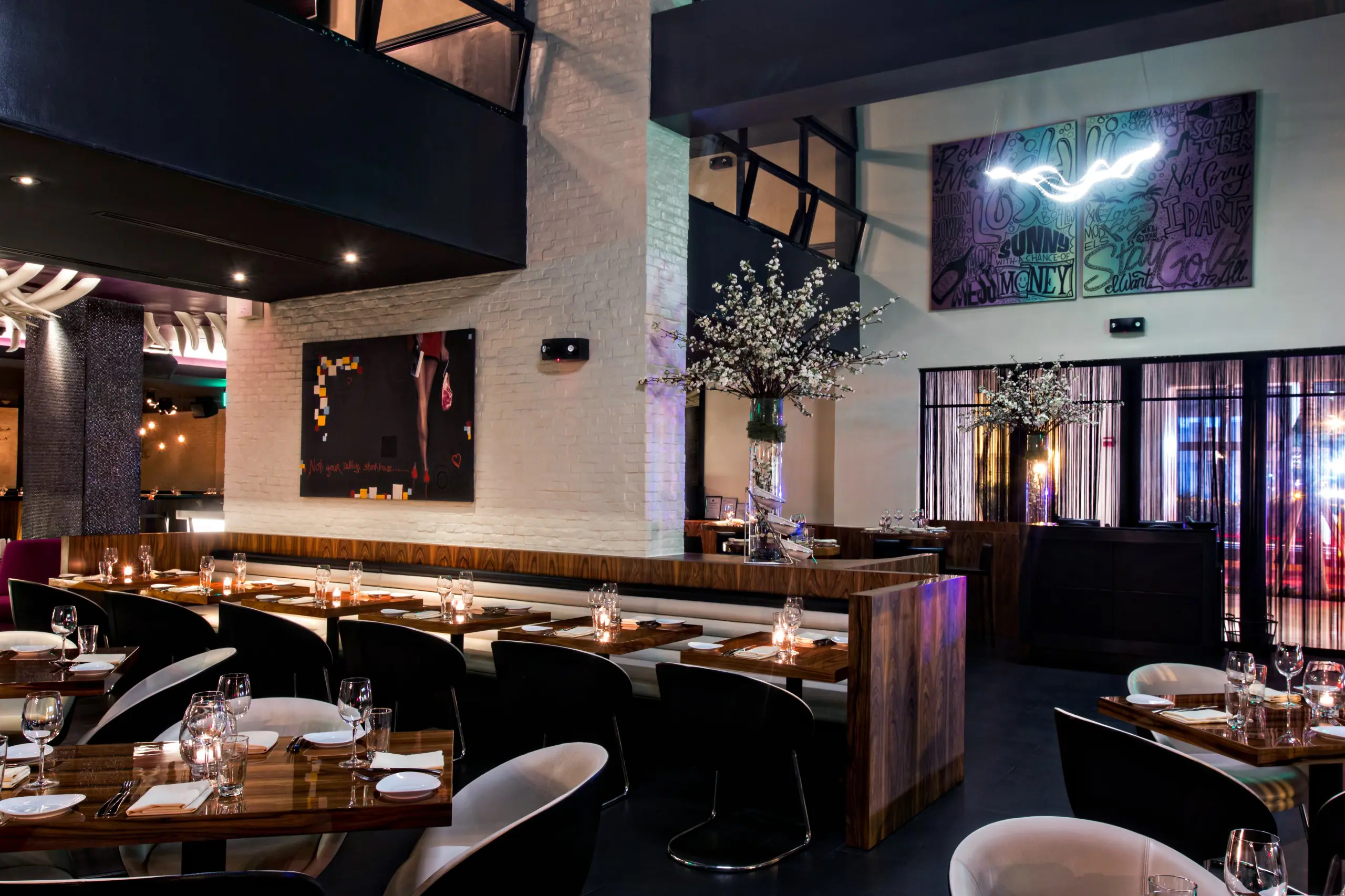 Interior of STK South Beach featuring dark wooden tables, dark chairs and ceiling, highlighted by a sculptural light fixture with looping neon like white tubing in the background. Tables are set with glassware and napkins, surrounded by contemporary white chairs with black legs.