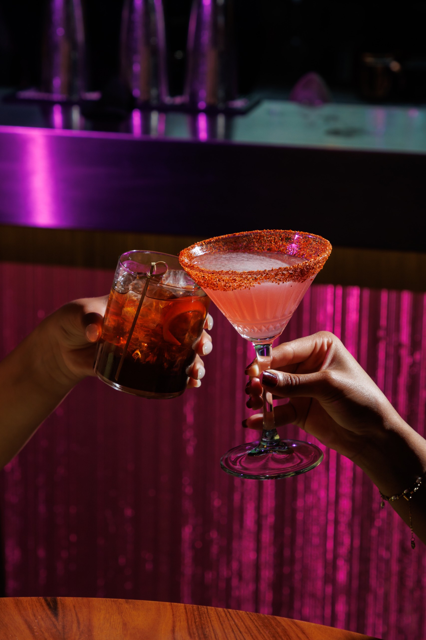 Two hands clinking cocktails at a bar: a dark drink in a rocks glass and a pink martini with a red sugar rim against a pink-lit backdrop.