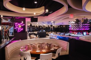 Interior of STK Charlotte restaurant featuring curved white banquette seating around a dark wooden table set with plates, glasses, and napkins. Overhead, the ceiling displays swirling white panels with built-in lighting. A neon pink sign reading "Dare to Be".