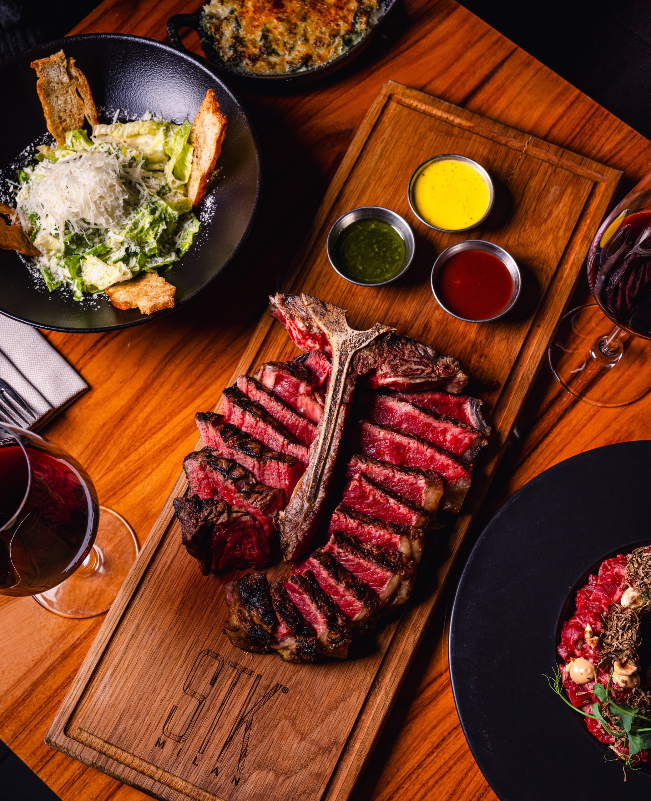 Overhead view of sliced bone-in steak on a wooden STK board with three dipping sauces, surrounded by Caesar salad, side dishes, and glasses of red wine on a wooden table.