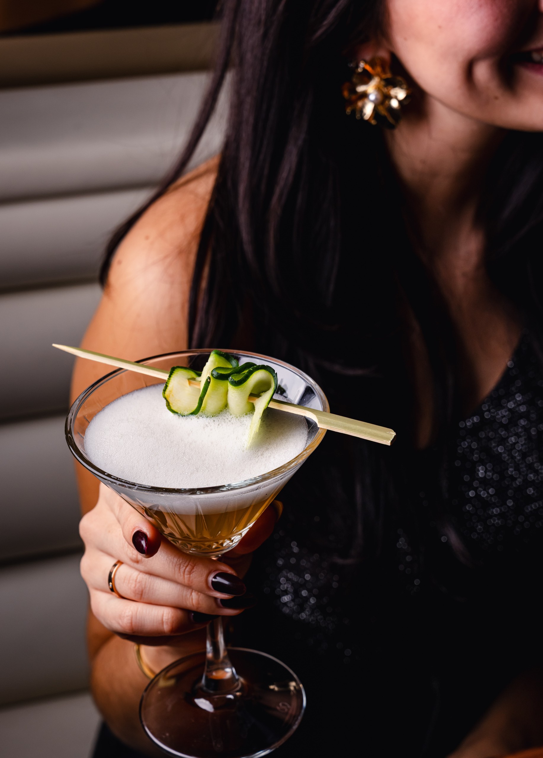 Woman holding a foamy cocktail in a martini glass garnished with a curled cucumber ribbon on a skewer in a dimly lit restaurant.
