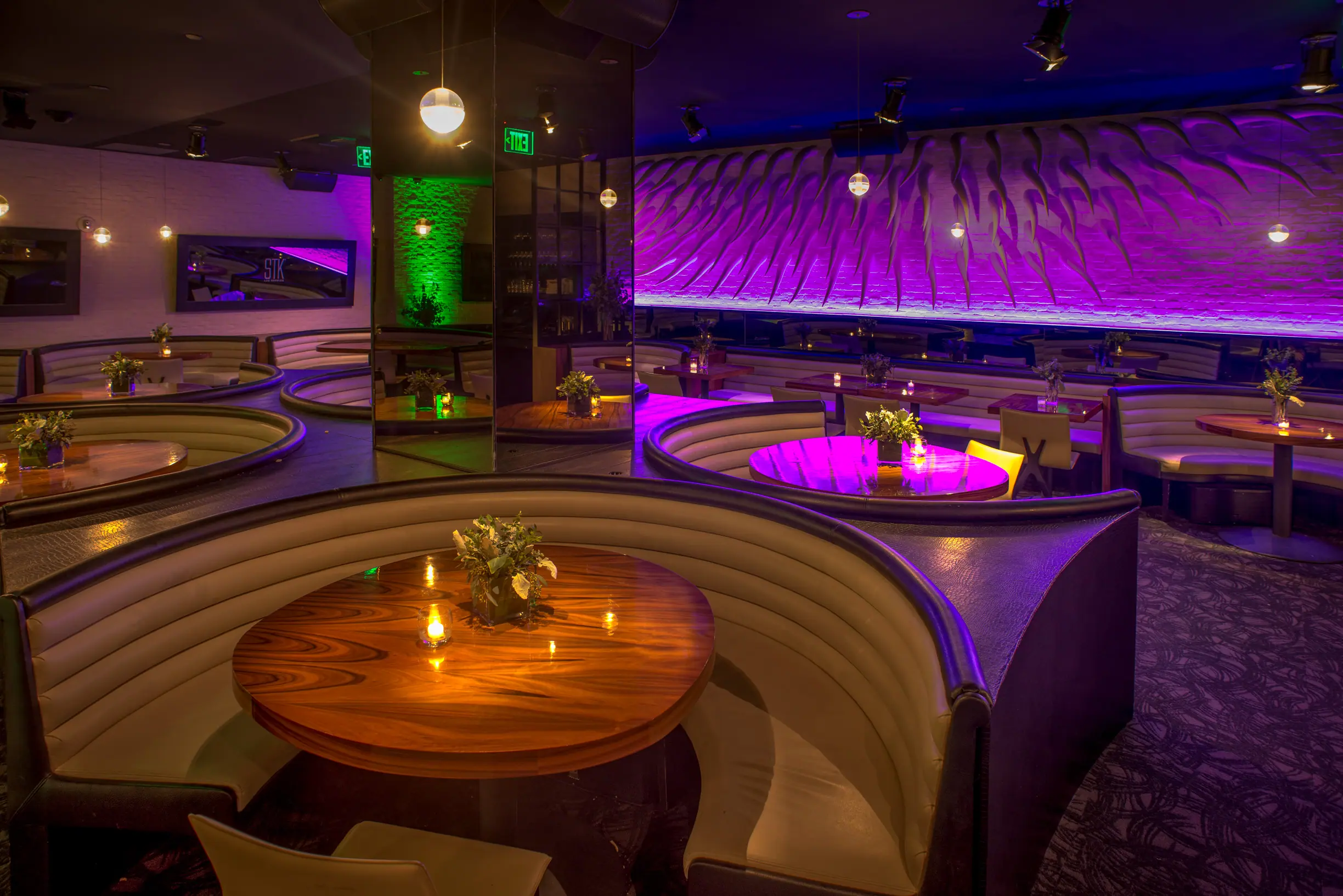 Interior of STK Los Angeles featuring curved booth seating upholstered in dark material. The booths are arranged in a semi-circular pattern around round wooden tables. Soft lighting and purple accent lighting is throughout.