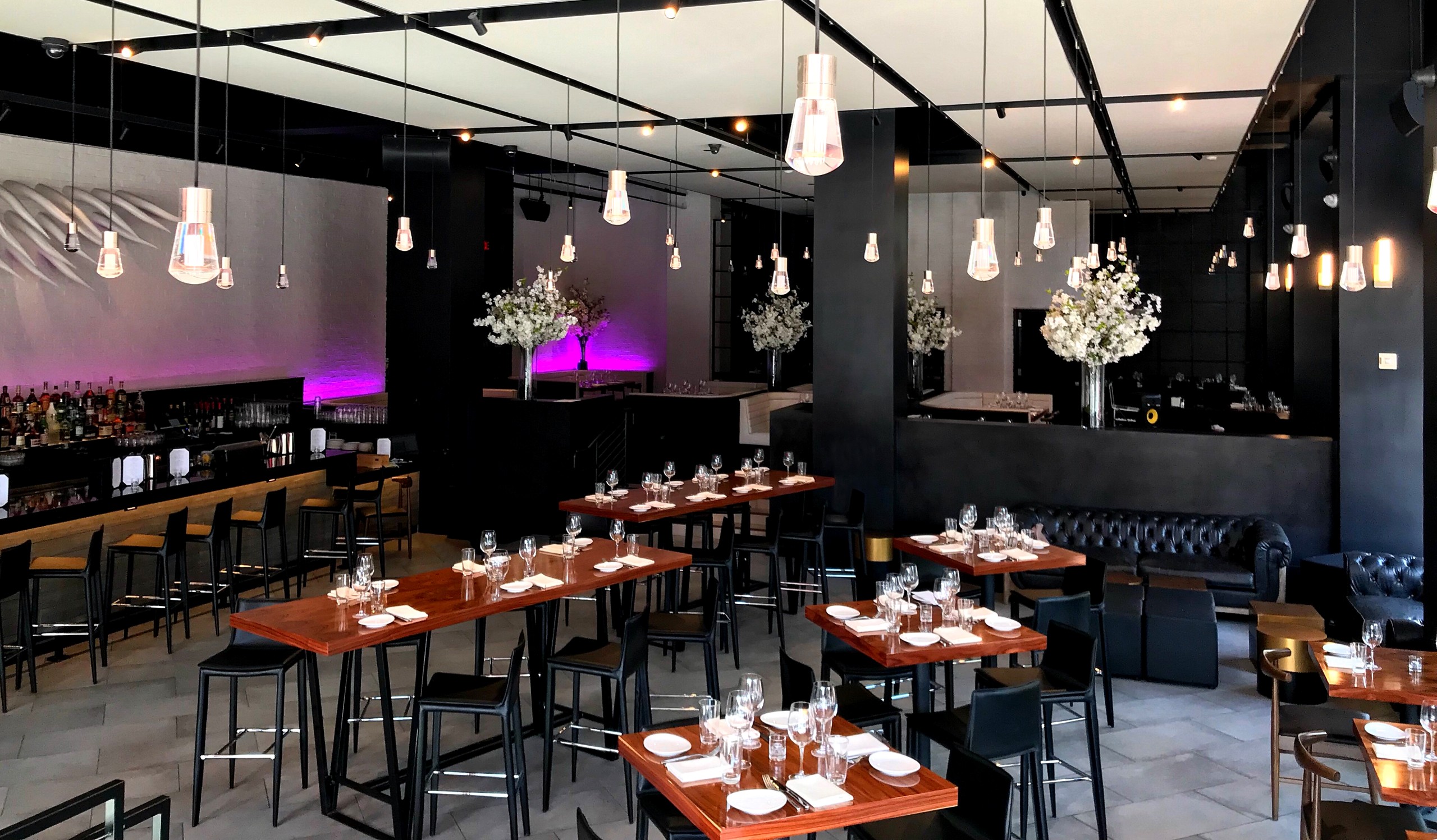 STK San Diego interior featuring dark walls and ceiling, with clusters of pendant lights and sculptural floral arrangements. Wooden tables are set for service, surrounded by a mix of black chairs and high stools. The bar area is illuminated with purple accent lighting.