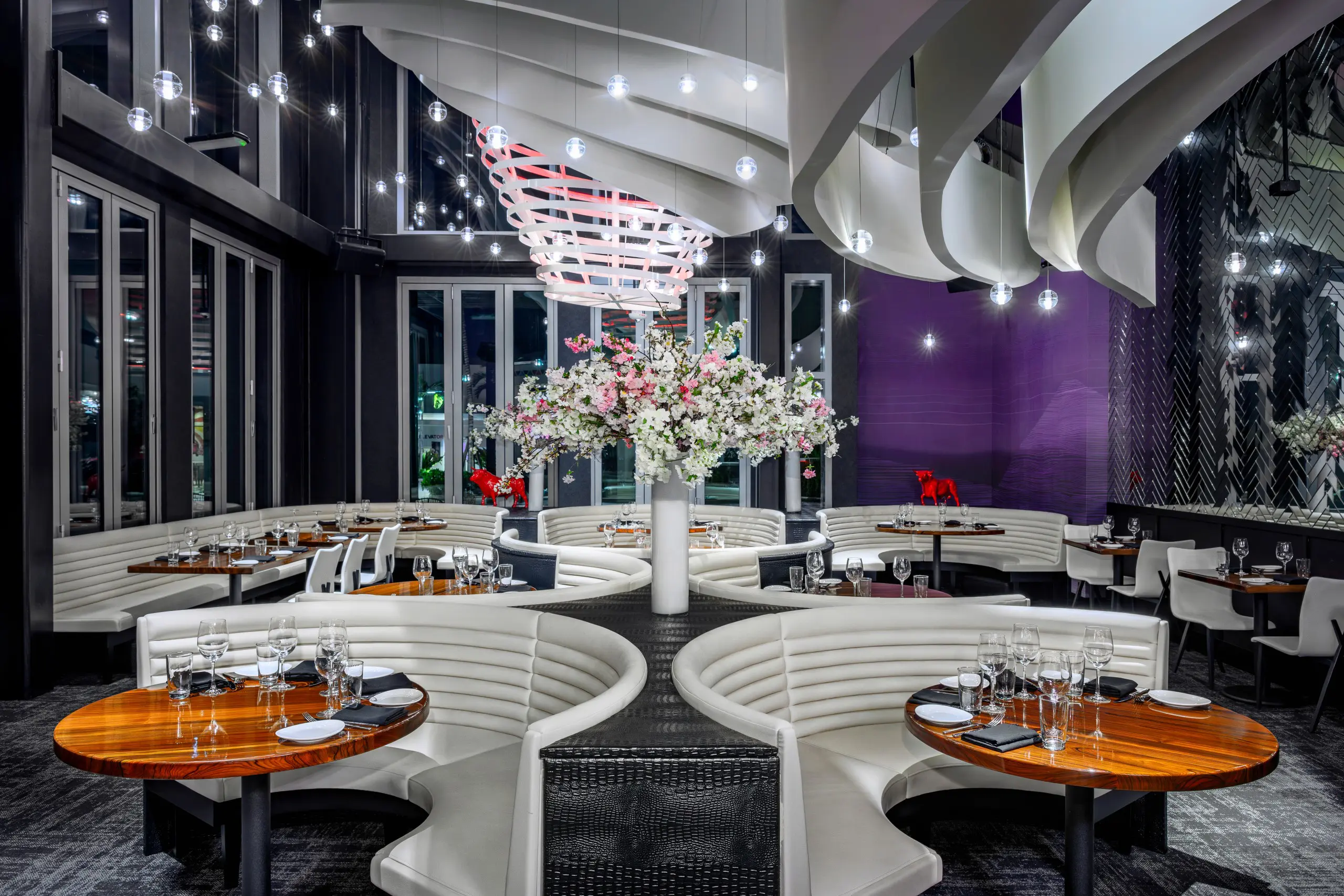 Interior of STK Aventura with curved white booth seating in semi circles around round wooden tables. A tall floral arrangement is centred beneath sculptural, wave like ceiling panels.