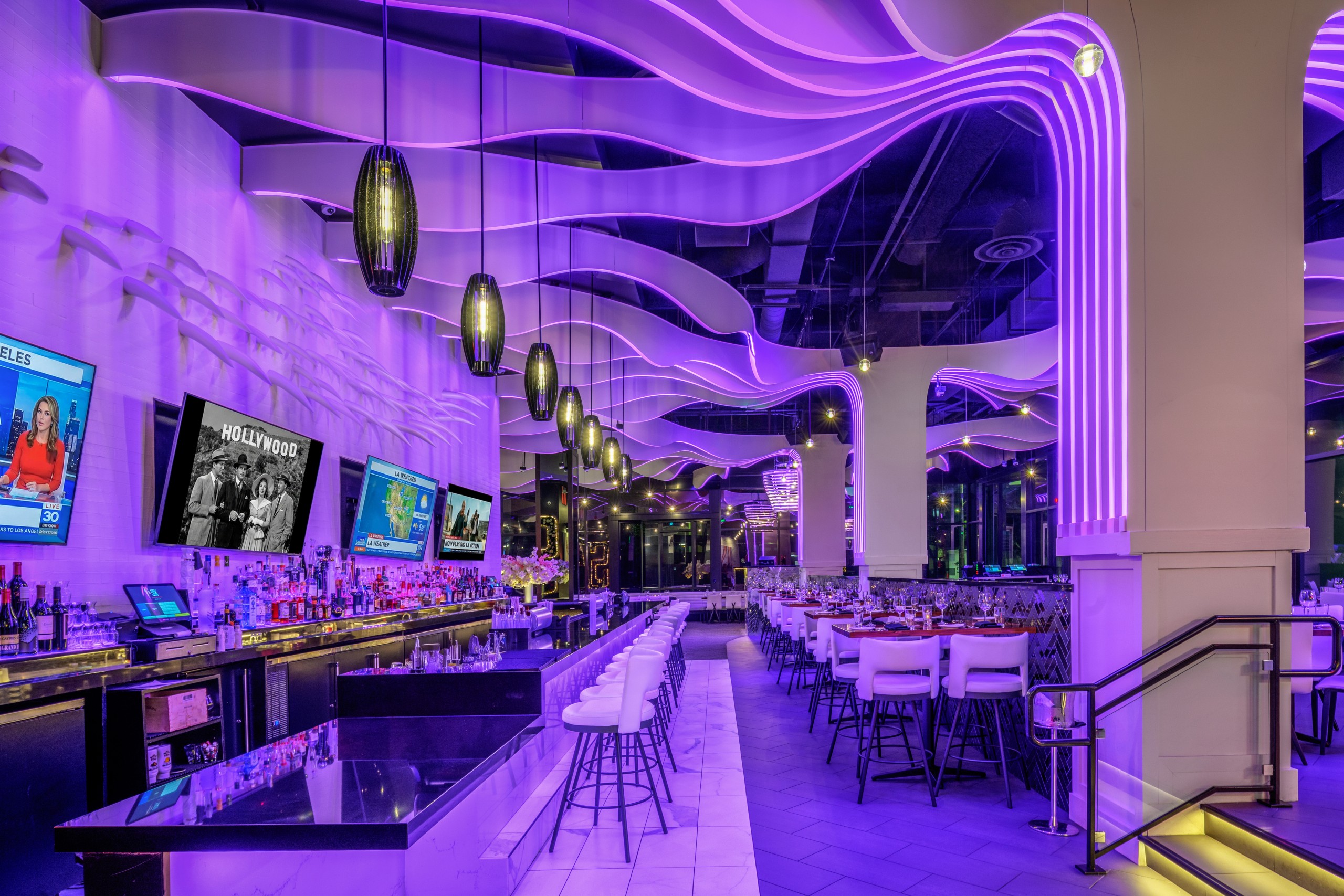 Vibrant bar and dining area with curved ceiling panels glowing purple, a long illuminated bar with high stools, wall-mounted TVs, and rows of white barstools and tables along the right side.