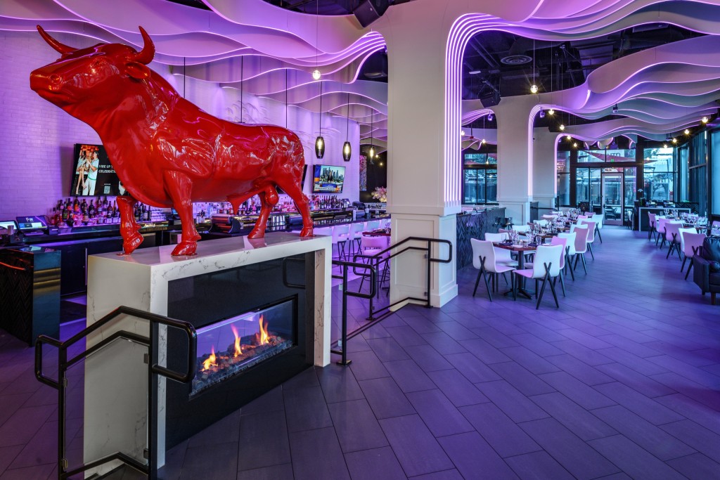 Modern bar and dining room with a large red bull sculpture above a glass fireplace, purple-lit wave ceiling, long bar on the left, and rows of white tables and chairs extending to the back.