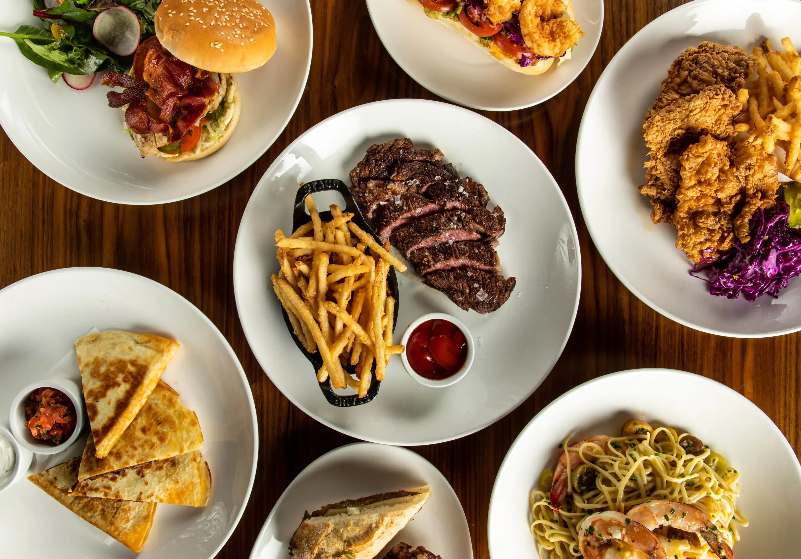 Meals on white plates viewed from above, including steak and fries, burgers and fried chicken