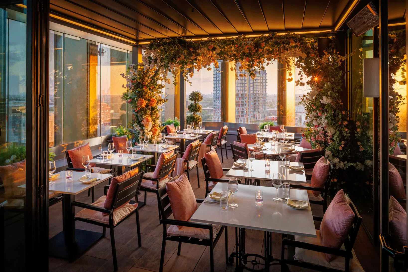 Rooftop restaurant dining area at sunset, with floor‑to‑ceiling windows, flower covered pillars, neatly set tables with glassware and napkins, cushioned chairs, and city skyline views in the background.
