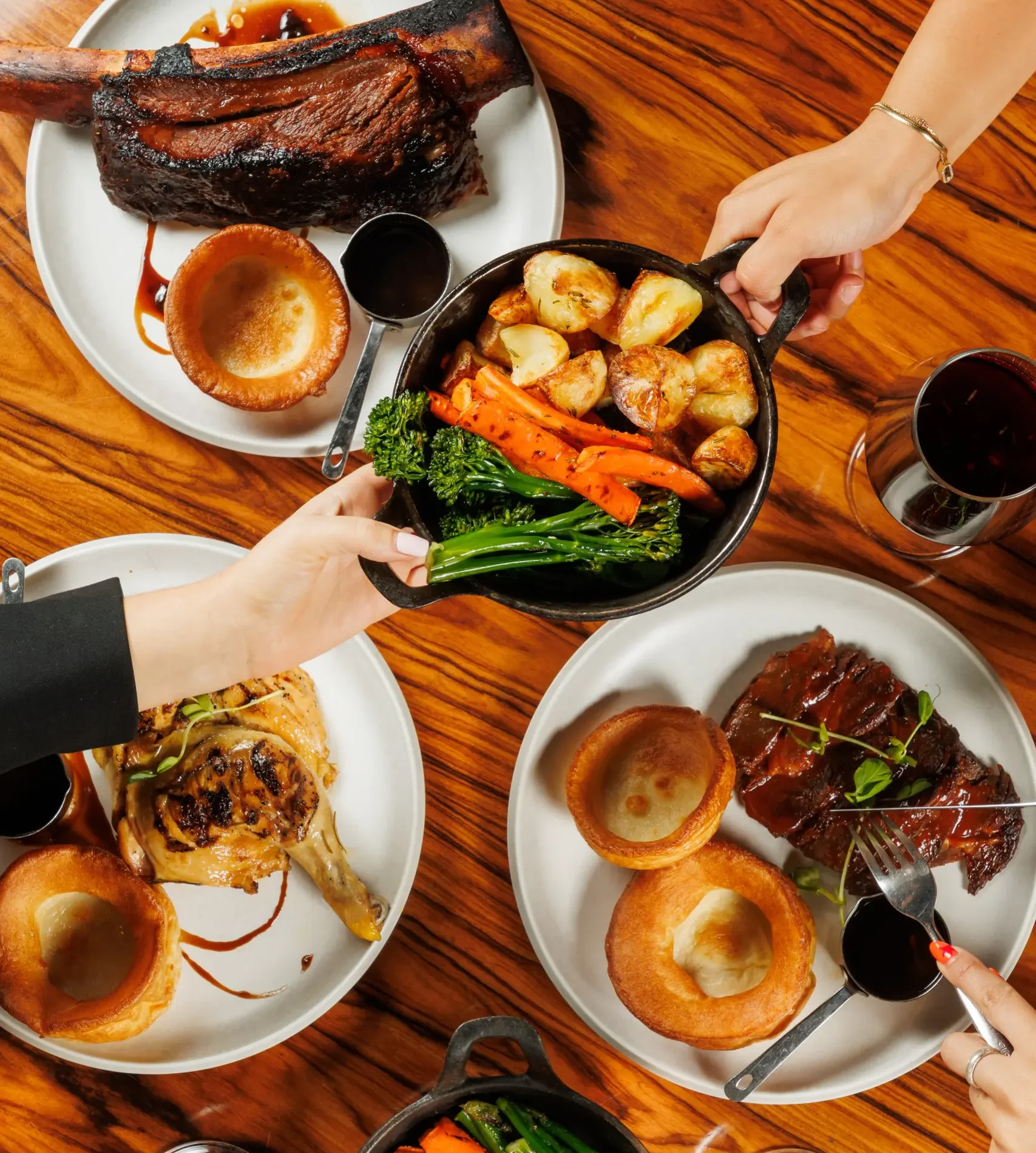 Top down view of a restaurant table with several plates of roast dinners, including Yorkshire puddings, sliced beef, roasted potatoes, carrots, green vegetables, and glasses of red wine, with diners’ hands serving and eating.