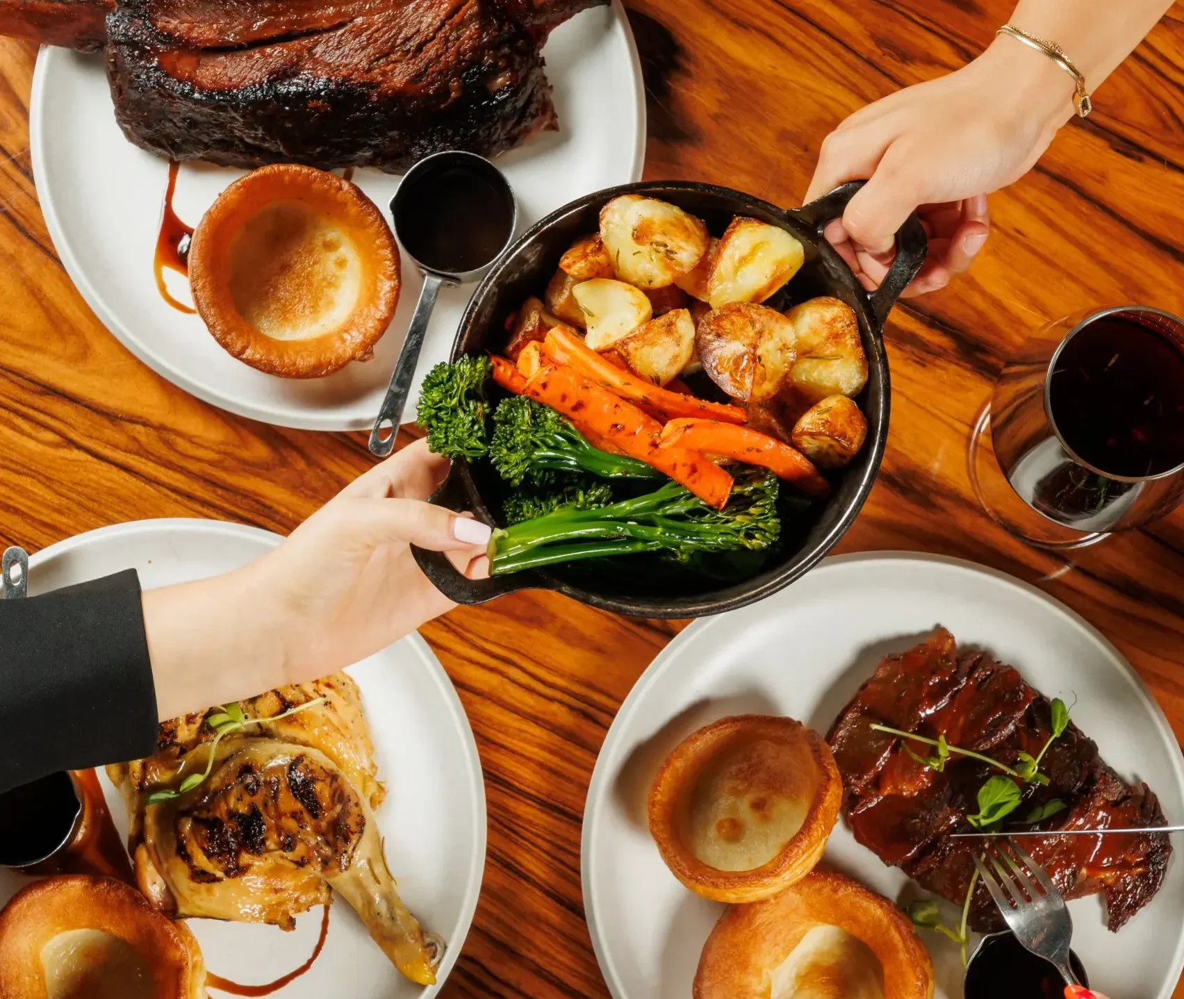 Top down view of a restaurant table with several plates of roast dinners, including Yorkshire puddings, sliced beef, roasted potatoes, carrots, green vegetables, and glasses of red wine, with diners’ hands serving and eating.