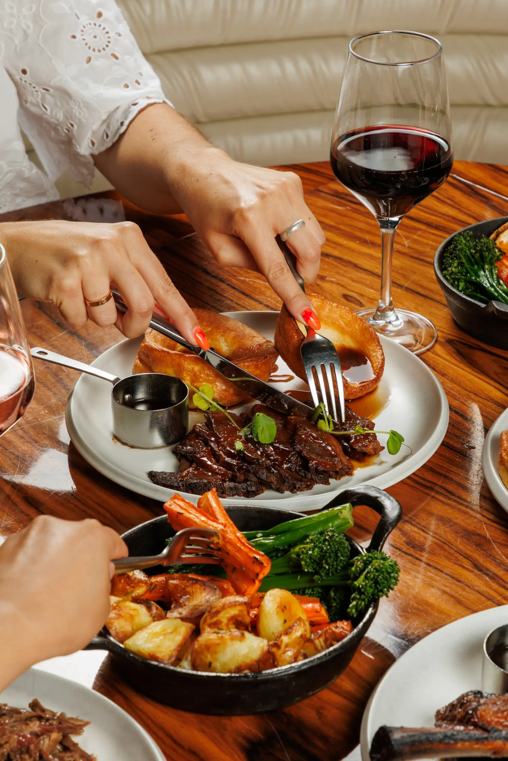 Close-up of people eating a roast beef dinner at a restaurant, with hands cutting meat and Yorkshire pudding on a white plate, side dishes of vegetables and potatoes, and glasses of red wine on a wooden table.
