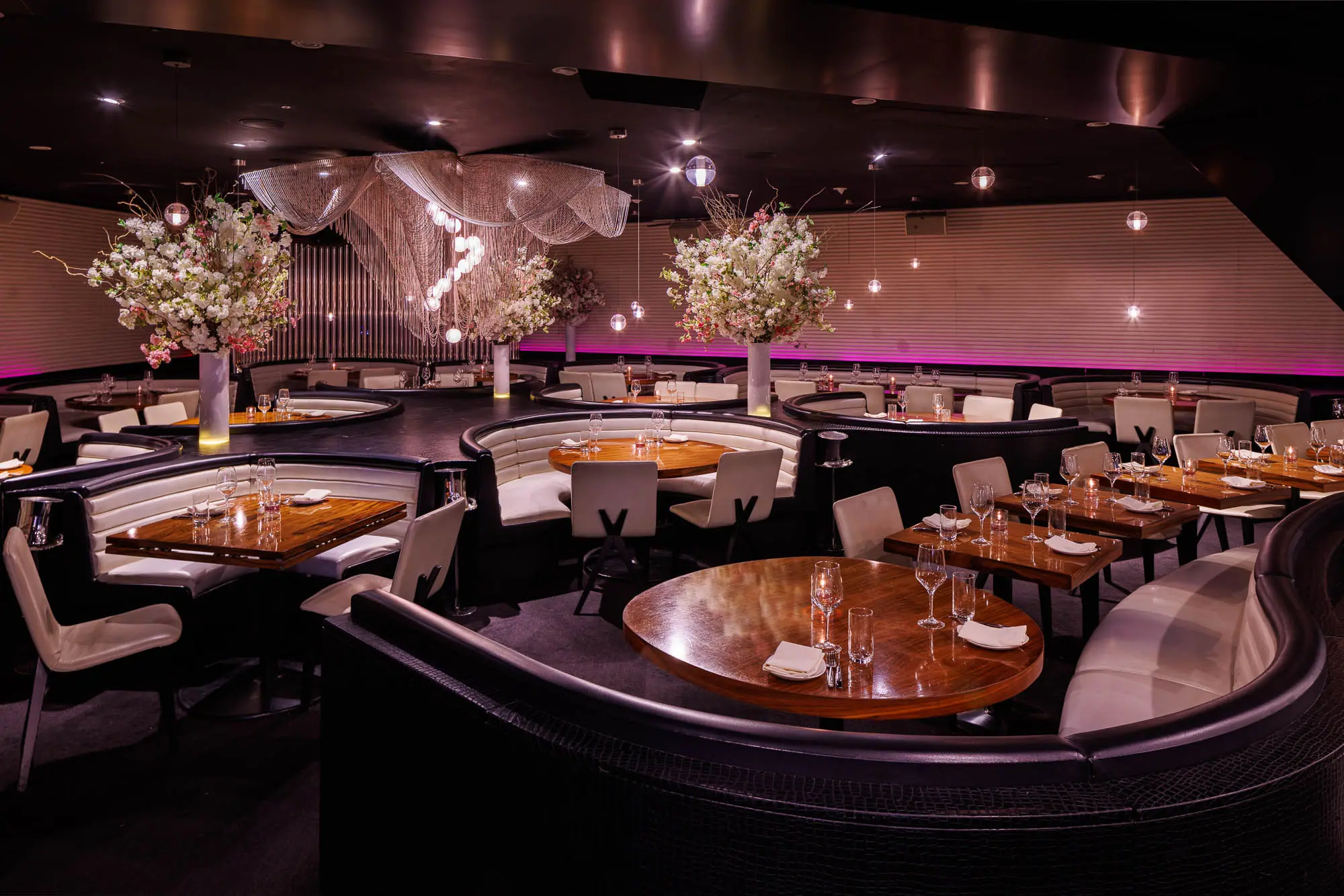 Restaurant interior with curved leather booths and wooden tables, set with glassware and napkins, featuring large floral centerpieces and dramatic hanging lights against a dark ceiling and softly lit walls.