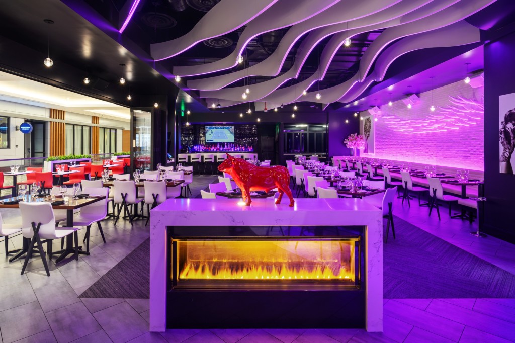 Contemporary dining room with purple-lit wave ceiling, a central marble fireplace topped by a red bull sculpture, and neatly set tables and chairs extending toward a bar with a TV at the back.