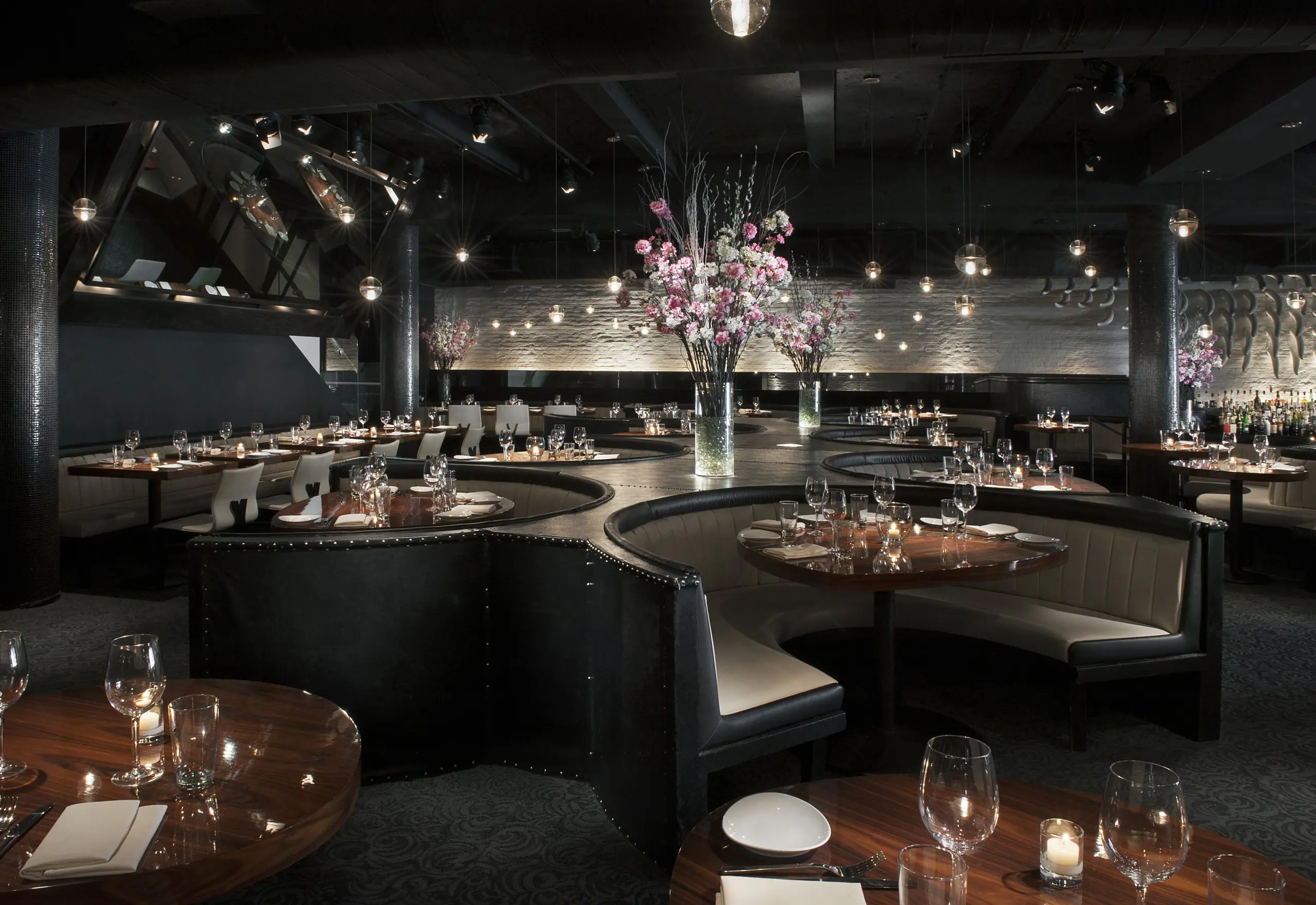 Interior shot showing curved black leather banquette seating, wood tables set with glassware and plates, floral centerpieces in tall vases, and pendant lighting.