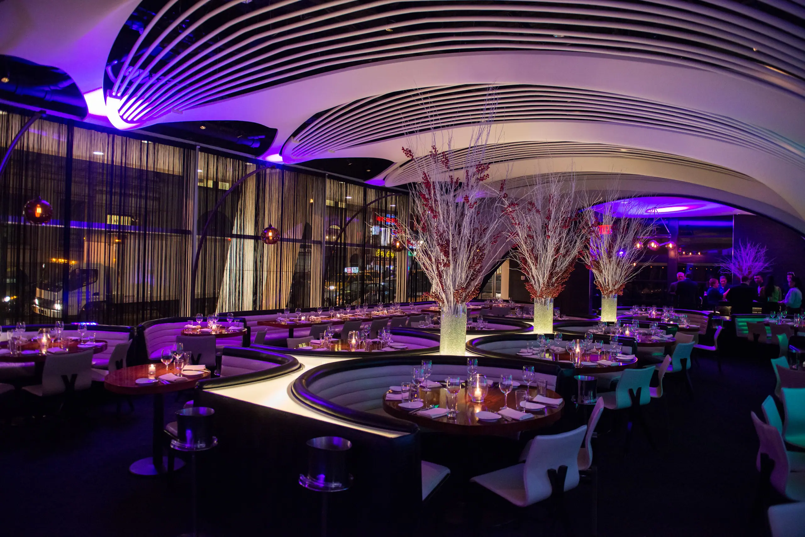 Interior shot with curved banquette seating, white branch centerpieces in illuminated vases. Tables are set, and ambient purple lighting under a sculptural ceiling.