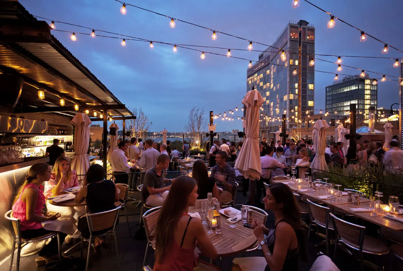 Lively rooftop restaurant at dusk, featuring string lights overhead, metal tables and chairs, groups of people dining and socializing.