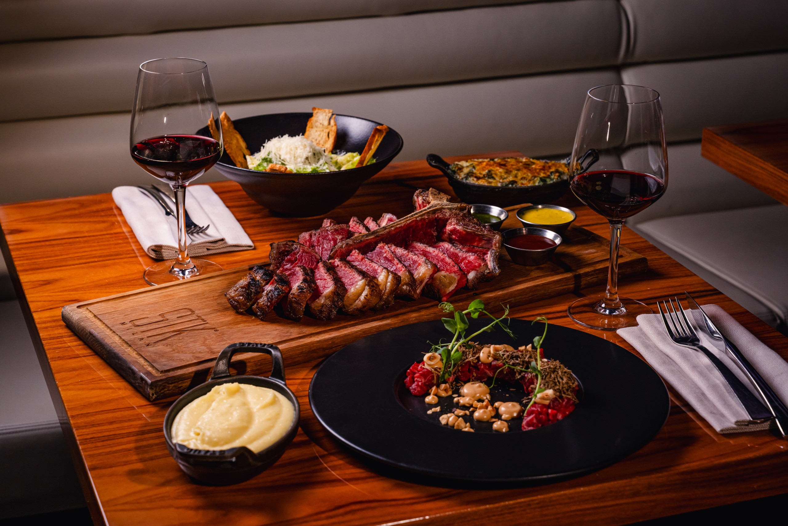 Plated steak dinner with sliced steak on a wooden board, sides, sauces and two glasses of red wine set on a polished restaurant table.