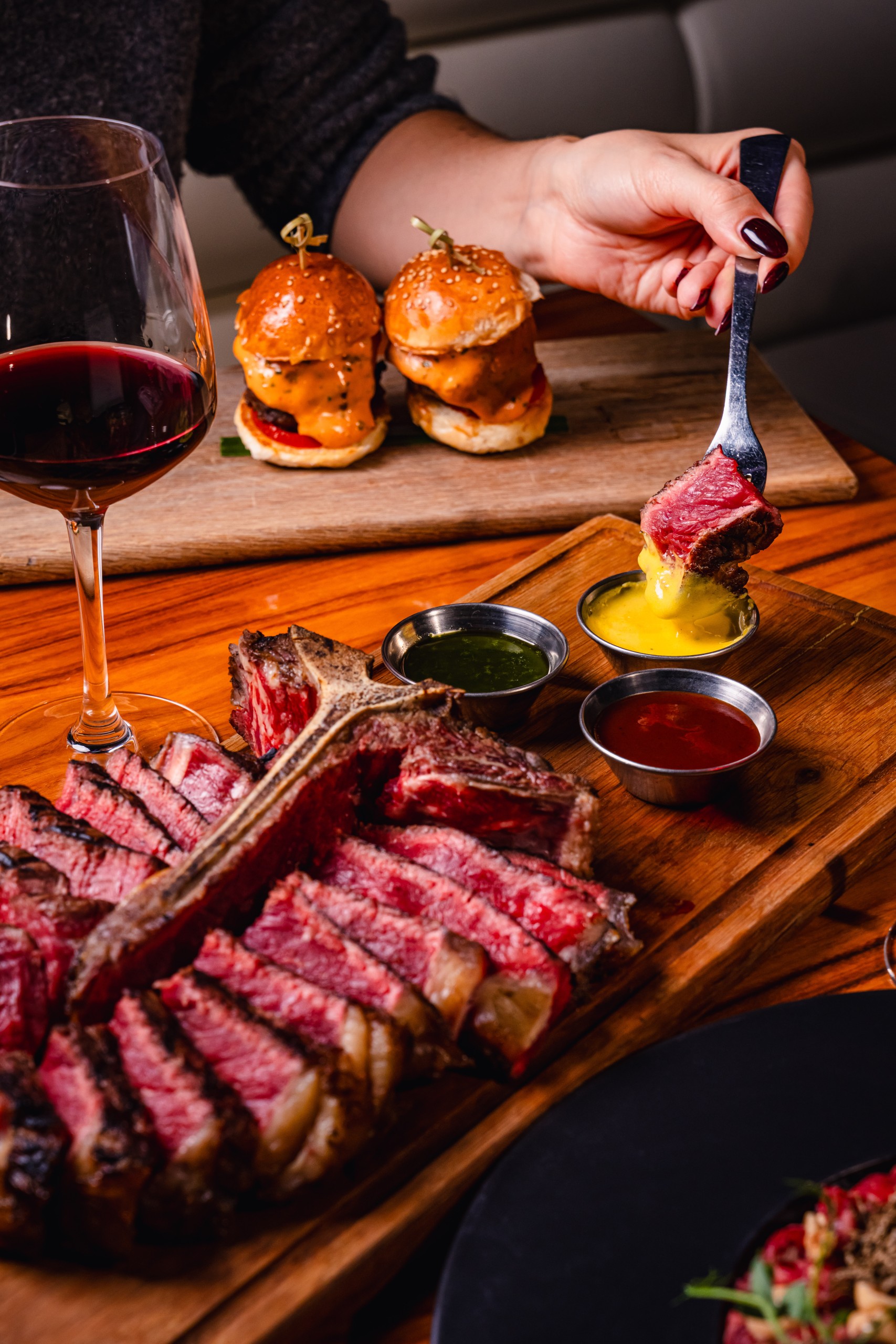 Steak dinner spread with sliced bone-in steak, dipping sauces, and a glass of red wine, while a hand dips a steak piece and two mini burgers sit on a wooden board in the background.