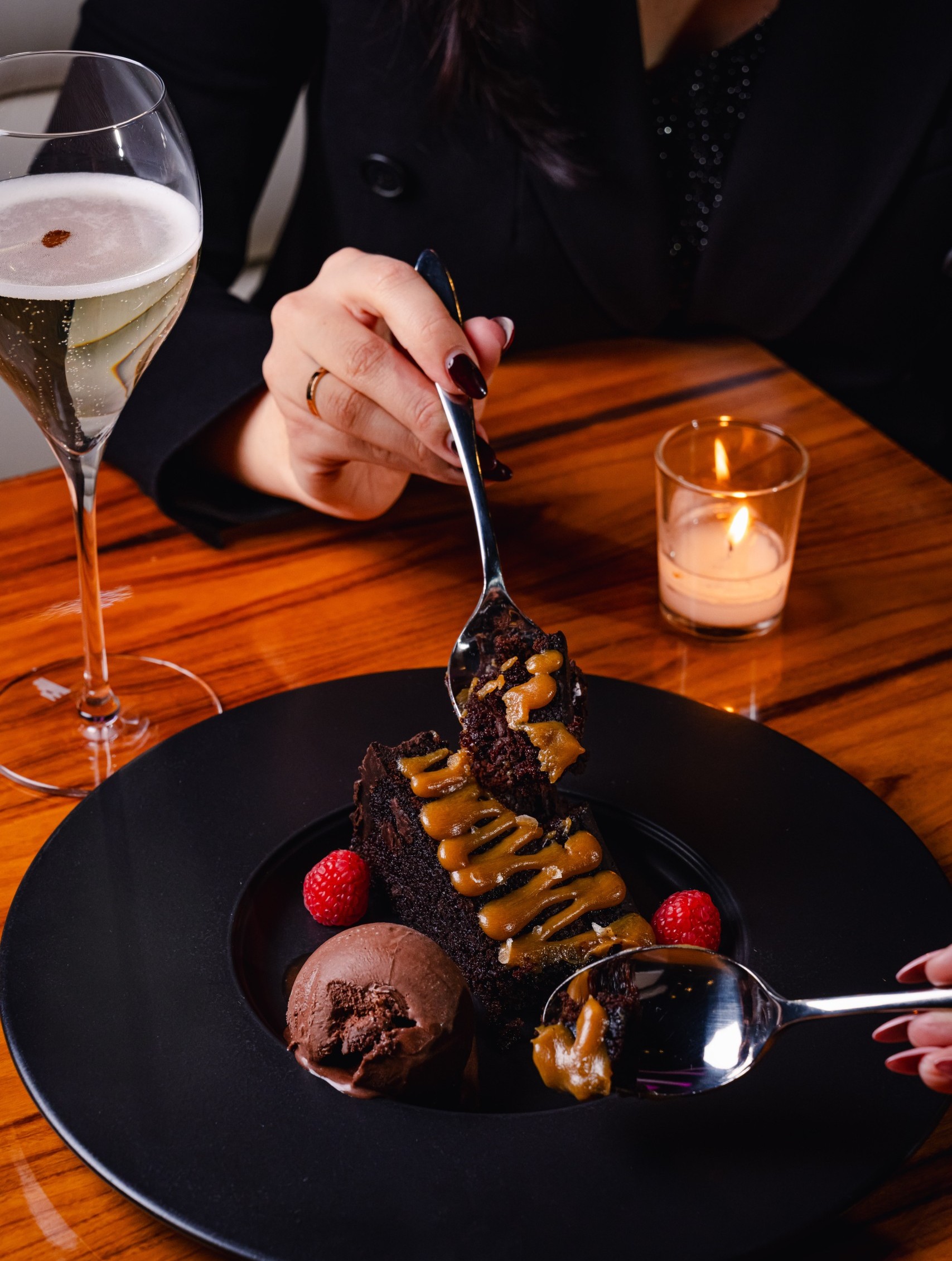 Hands sharing a plate of chocolate cake with caramel drizzle, chocolate ice cream, and raspberries beside a glass of sparkling wine and a lit candle on a wooden table.