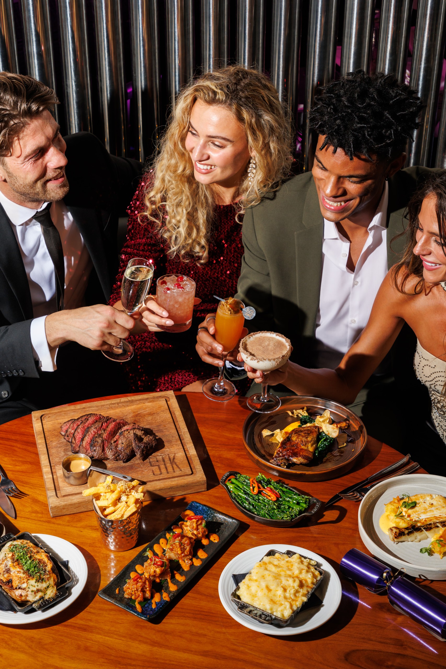 Group of friends dressed for a night out, clinking cocktails over a table filled with steak, sides and plated dishes at a restaurant.