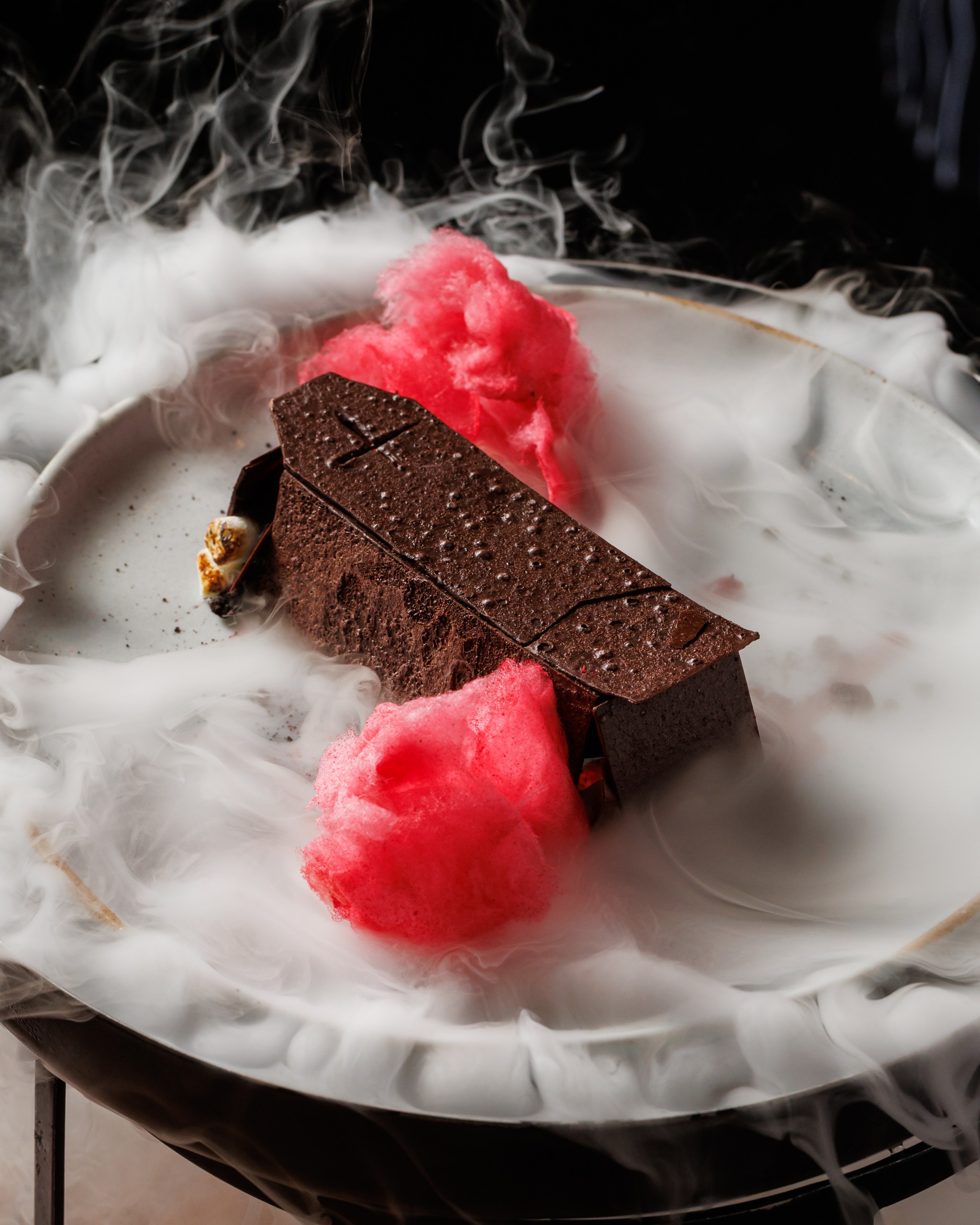 Plated chocolate coffin-shaped dessert with pink cotton candy and dramatic dry-ice fog swirling around the dish.