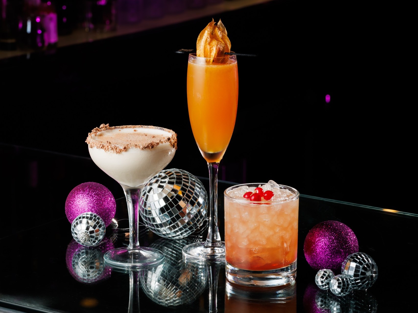 Three festive cocktails on a reflective bar top surrounded by purple ornaments and silver disco balls.