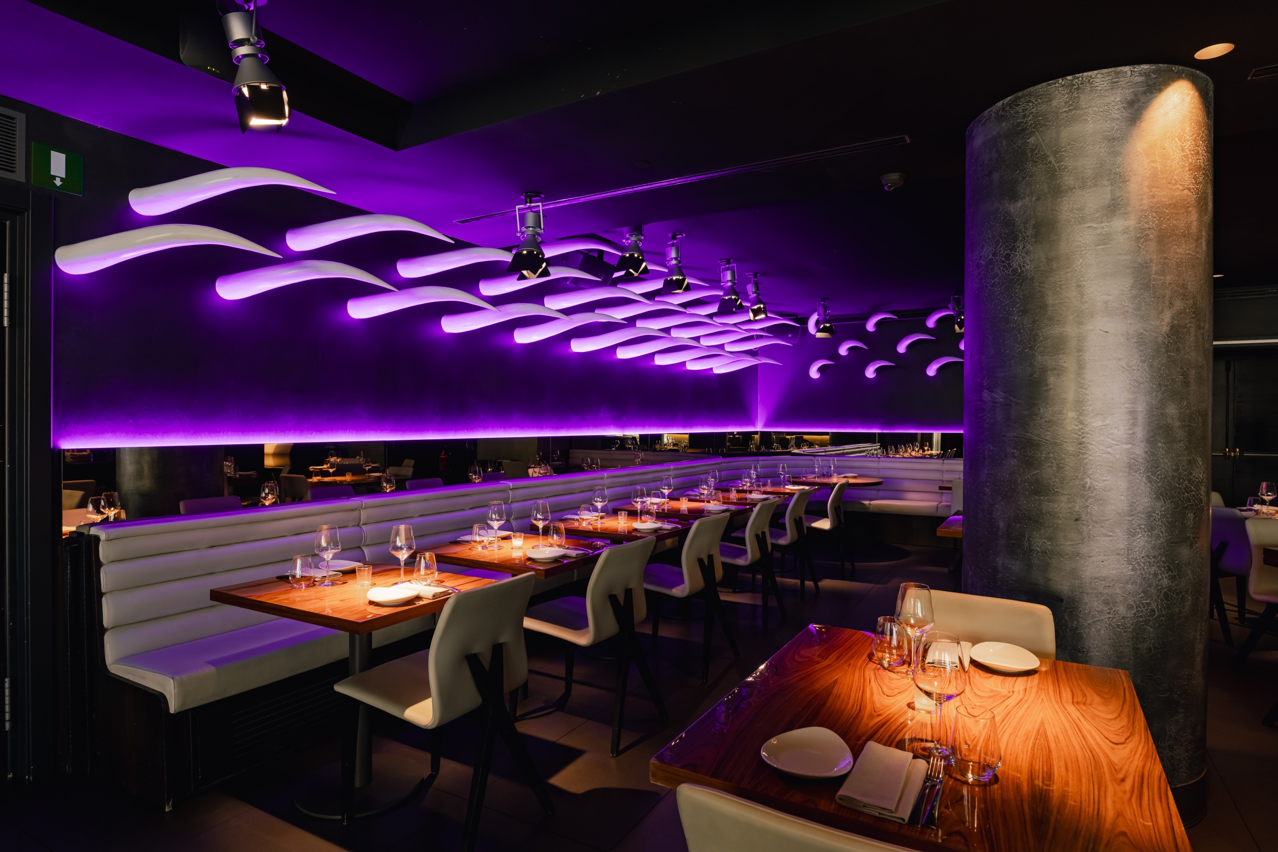 Moody dining area with white banquette seating, wood tables set for service, and sculpted ceiling lights casting purple glow along the length of the room.
