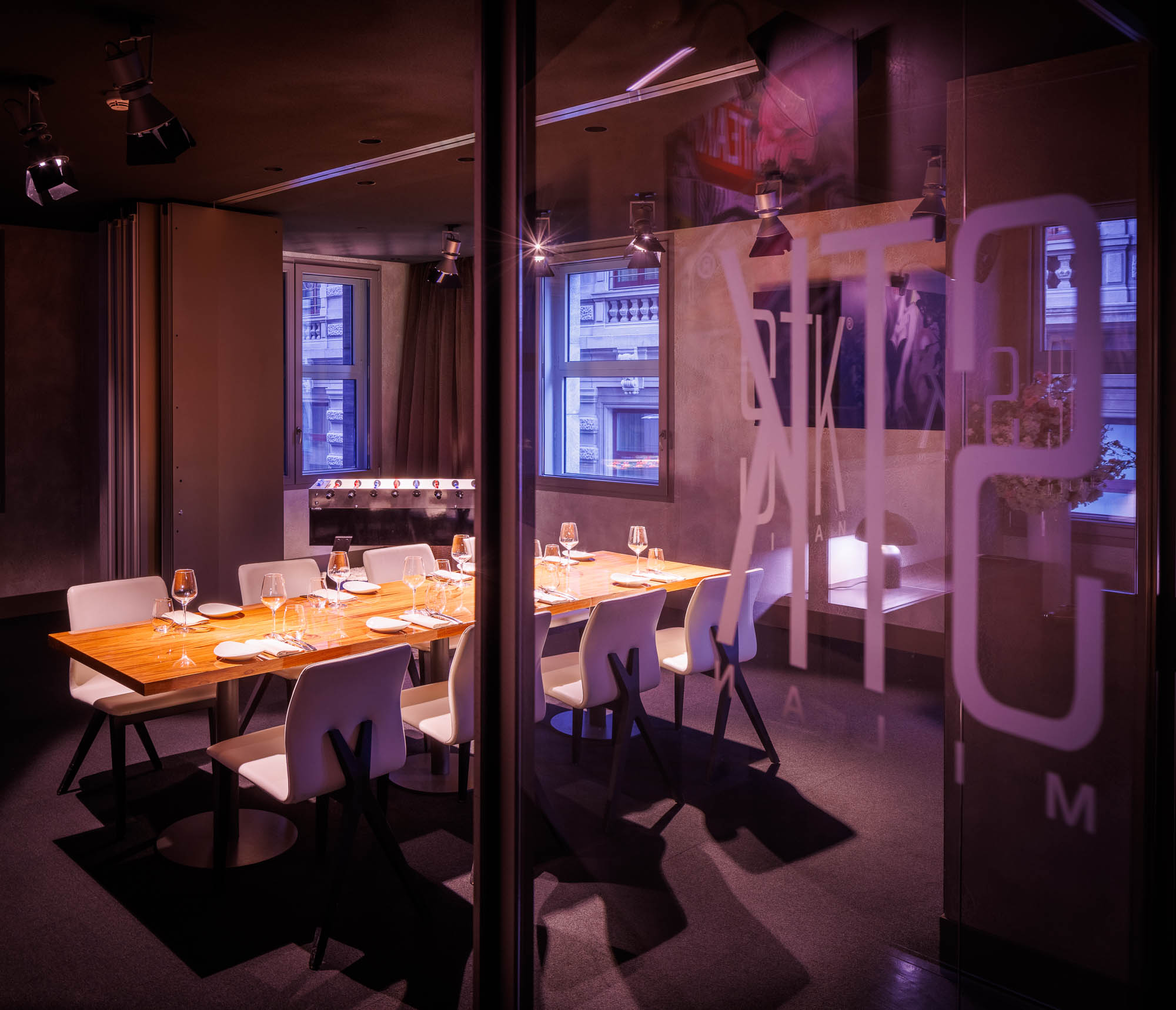 Close up of glass enclosed private dining room with a long wooden table, white chairs, purple lighting, and large STK Milan branding on the glass wall.