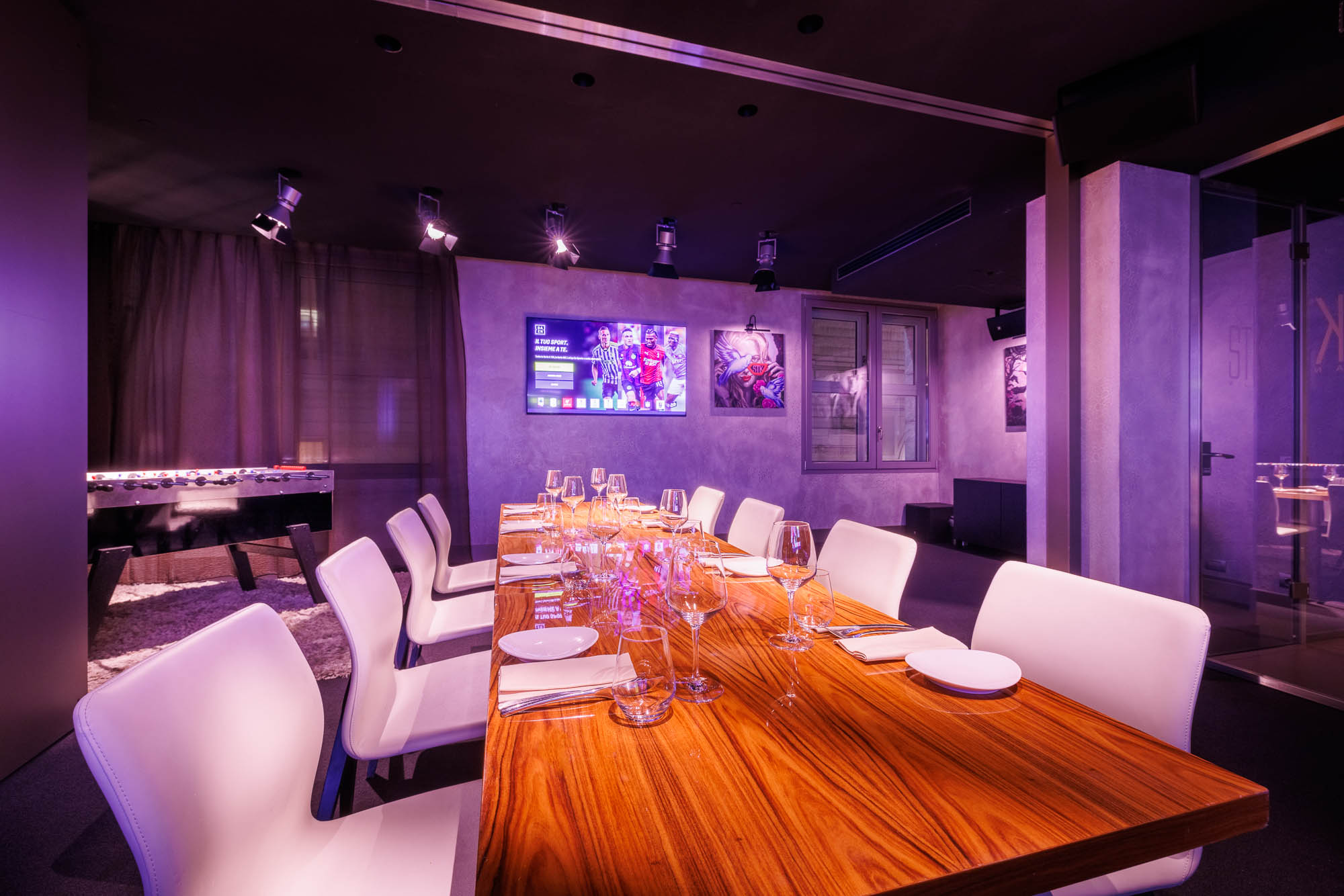 Private dining room with a long wooden table set with glassware and white chairs, purple ambient lighting, and a foosball table in the background.