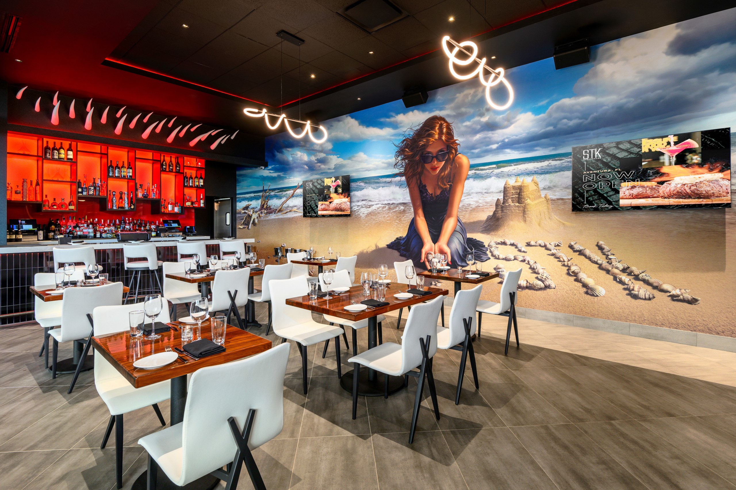 Vibrant restaurant dining room with white chairs, wood tables set for service, a glowing red backlit bar, and a large beach mural featuring a woman sitting by a sandcastle on the wall.