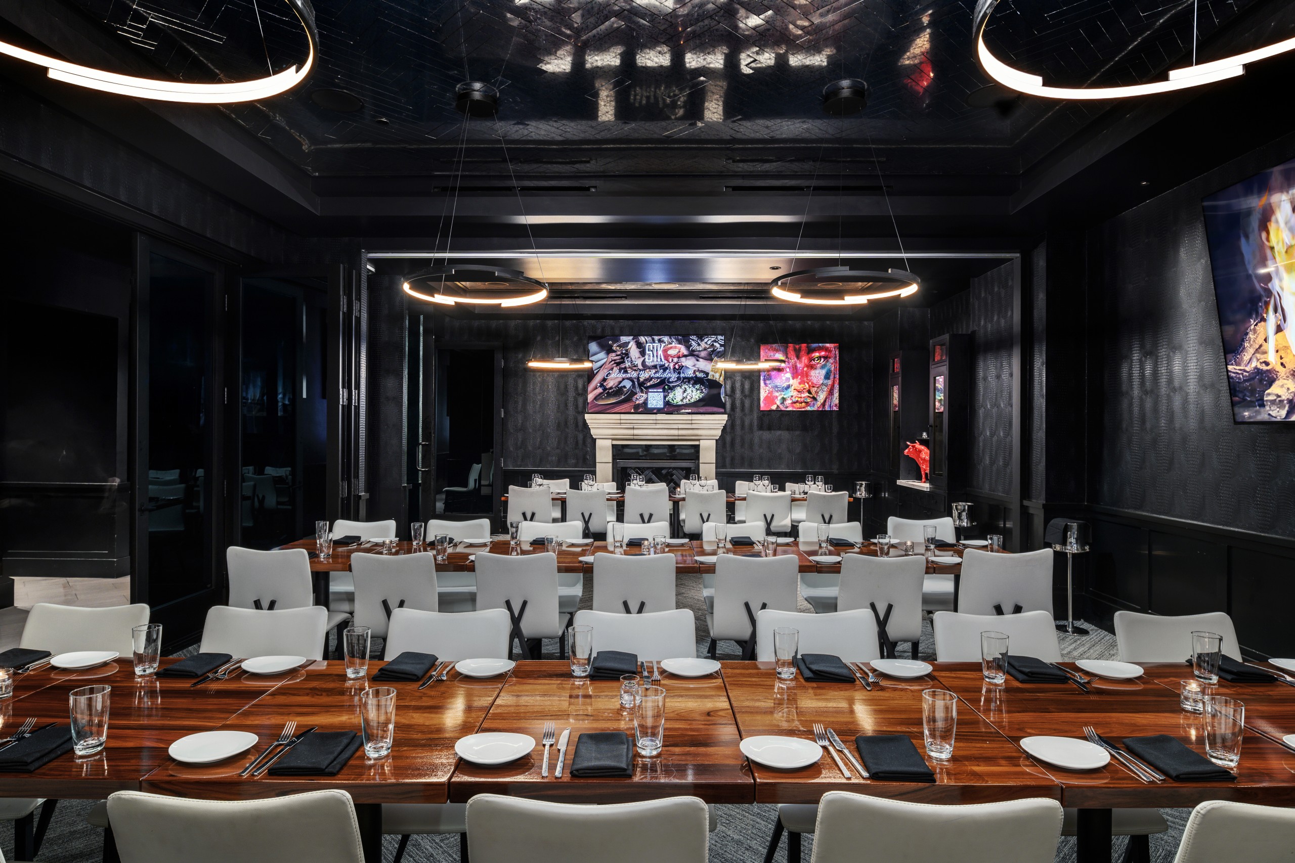 Private dining room with long wood tables set for service, white chairs, dark textured walls, and circular modern light fixtures hanging from the ceiling.