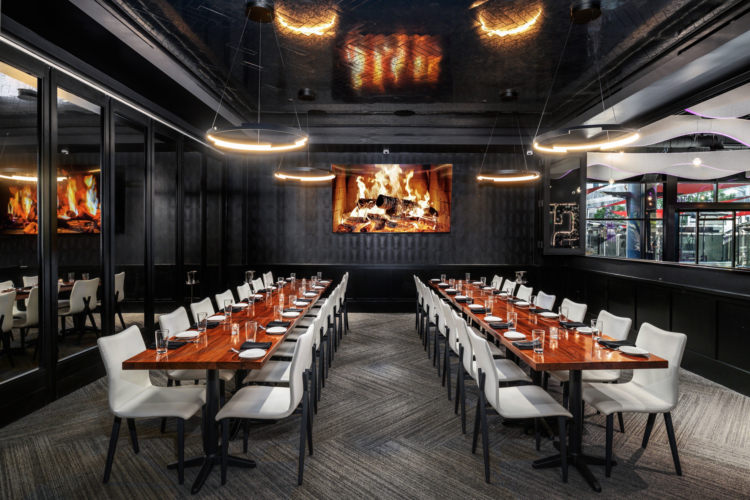 Long private dining room with two parallel wood tables set for service, white chairs, dark walls, circular modern light fixtures, and a large fireplace style video screen showing burning logs.