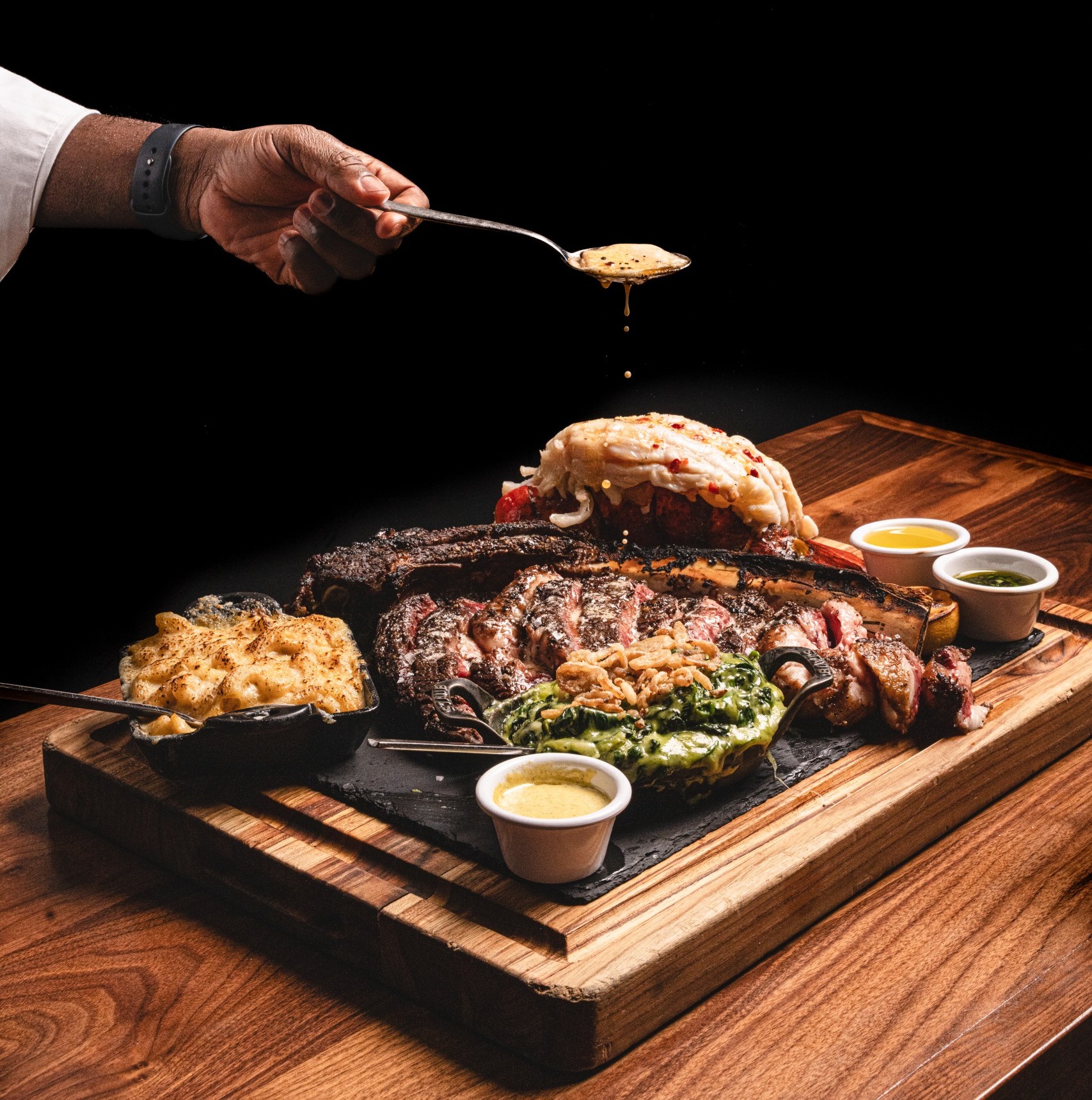 Close up of a wooden board loaded with grilled steak, lobster, sides, and dipping sauces, as a chef’s hands finish the dish by torching and spooning sauce over the meat against a black background.