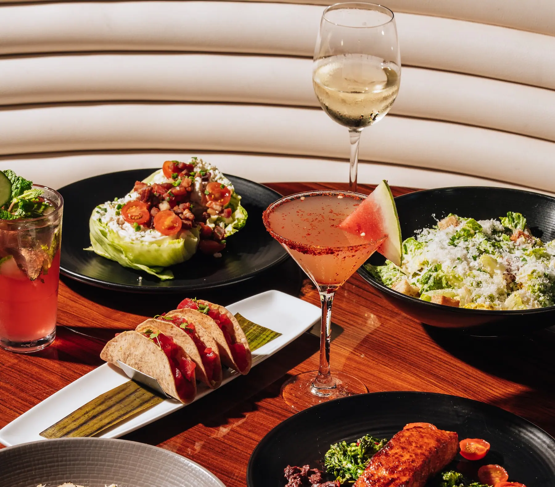 Tabletop spread of restaurant dishes, including a wedge salad topped with cherry tomatoes, a Caesar-style salad, three small tacos on a long white plate, a glazed salmon entrée with vegetables, and two cocktails. Set on a polished wooden table in front of a curved, cream-colored banquette.
